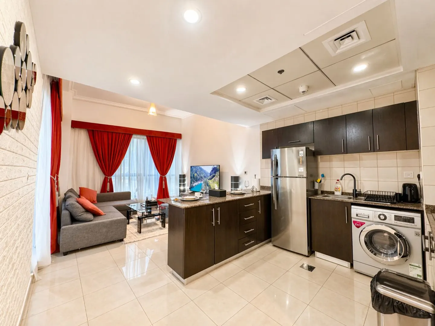 A modern apartment interior with a gray sofa, red curtains, and a kitchen area with stainless steel appliances.