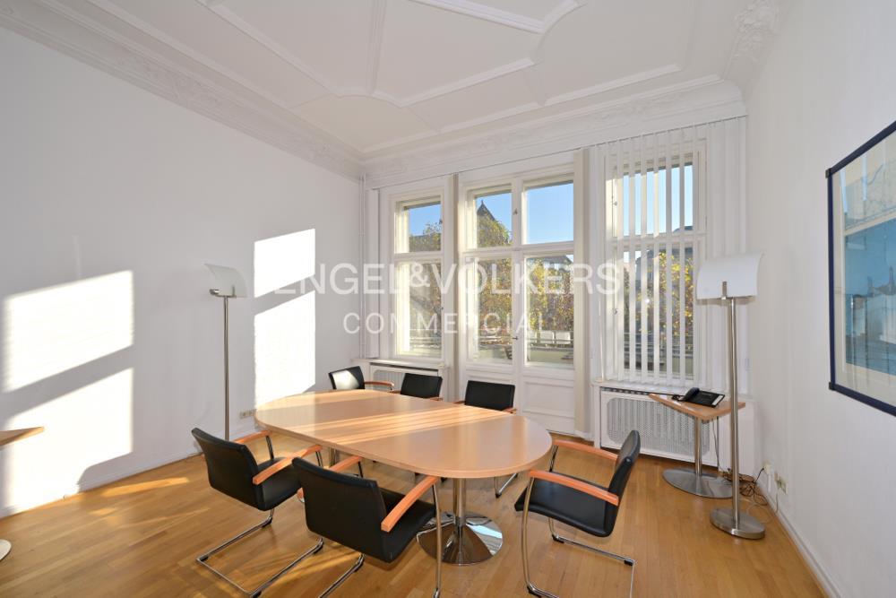 Bright meeting room with a light wood table, black chairs, wood floors, and large windows with white blinds.