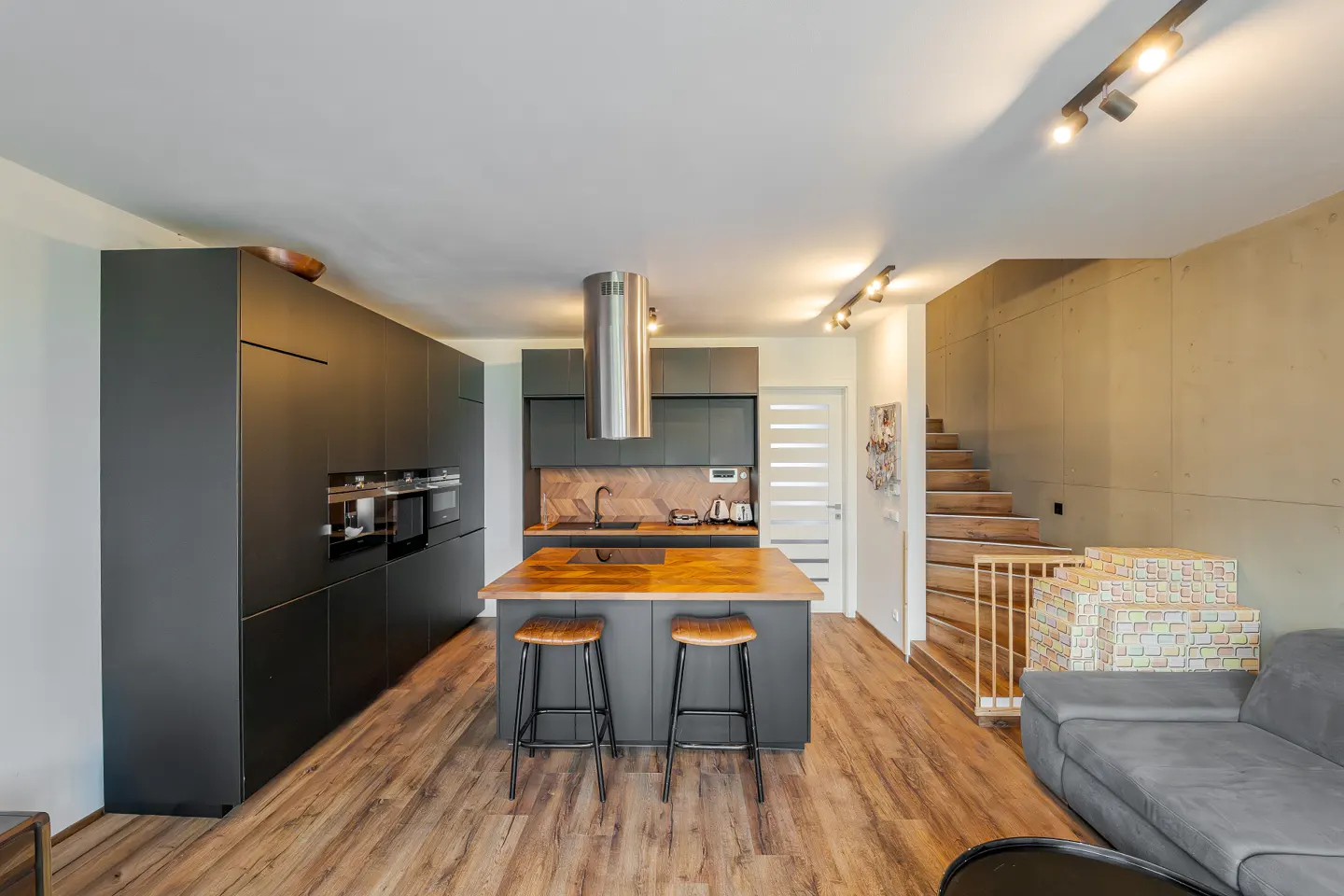 Modern kitchen with dark cabinets, wood floors, and island with stools. Stairs lead to the upper level. Gray sofa is visible in the foreground.