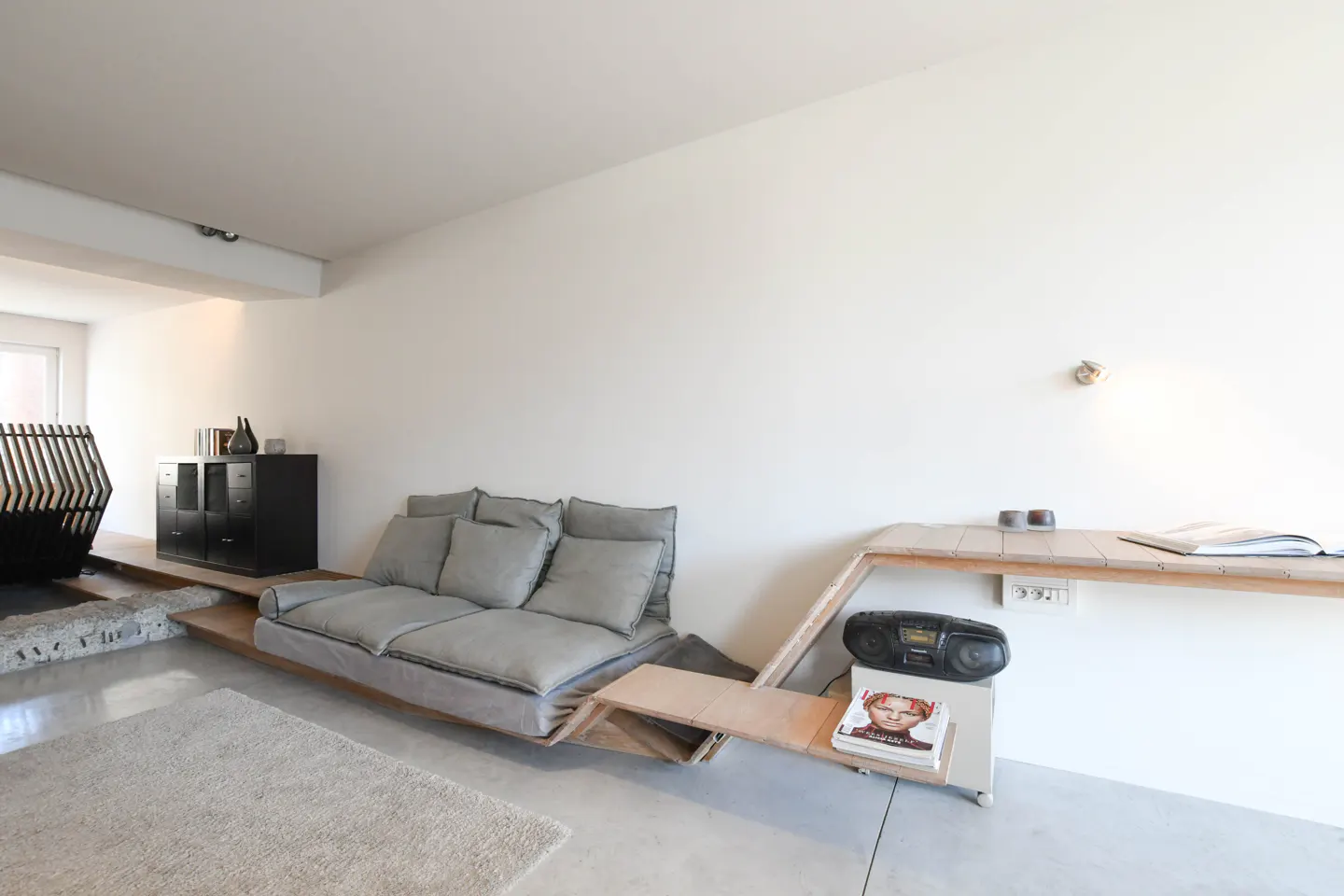 Minimalist living room with a gray sofa, wooden platform, black cabinet, and white walls. A boombox and books sit on a shelf.