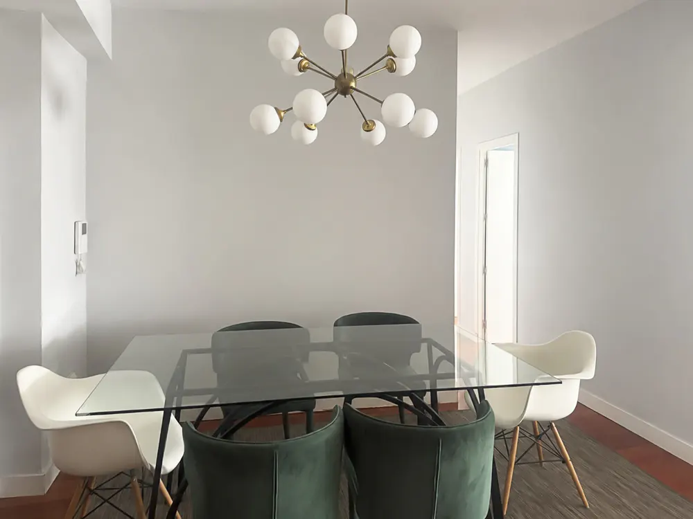 Modern dining room with a glass table, two white molded chairs, and four dark green velvet chairs under a globe chandelier.