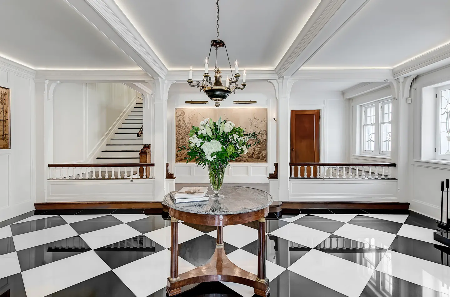 Elegant foyer with black and white checkered floor. A round table holds flowers and books. Stairs and a chandelier add to the luxurious feel.