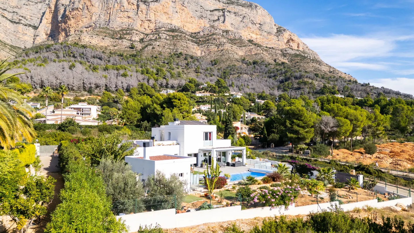 Modern white villa with a pool, surrounded by lush greenery and a mountain backdrop on a sunny day.