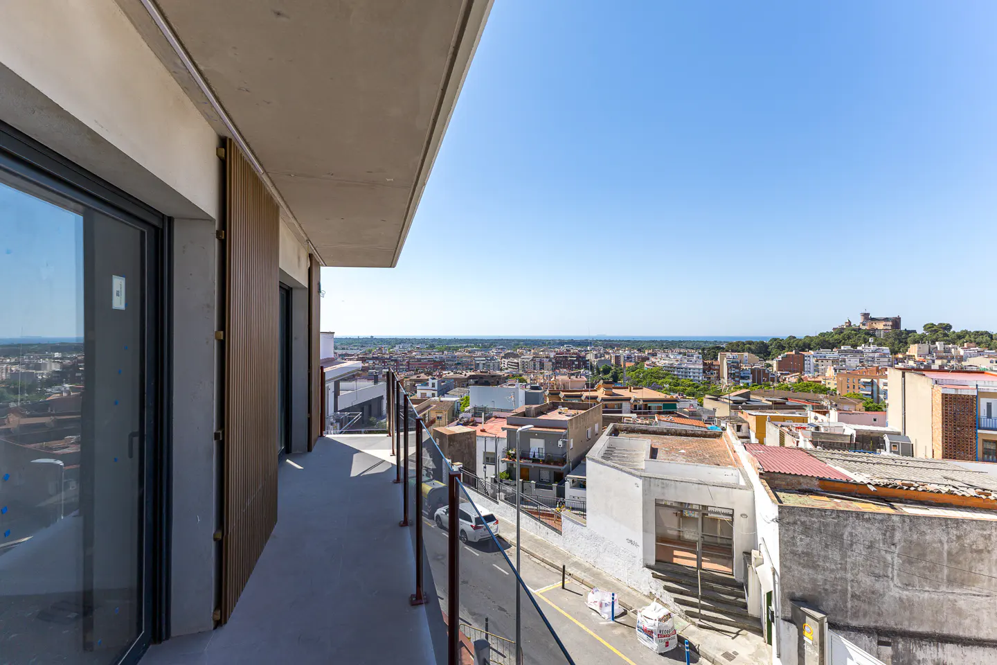 Modern balcony with glass railing overlooking a sunny town with a distant castle under a clear blue sky.