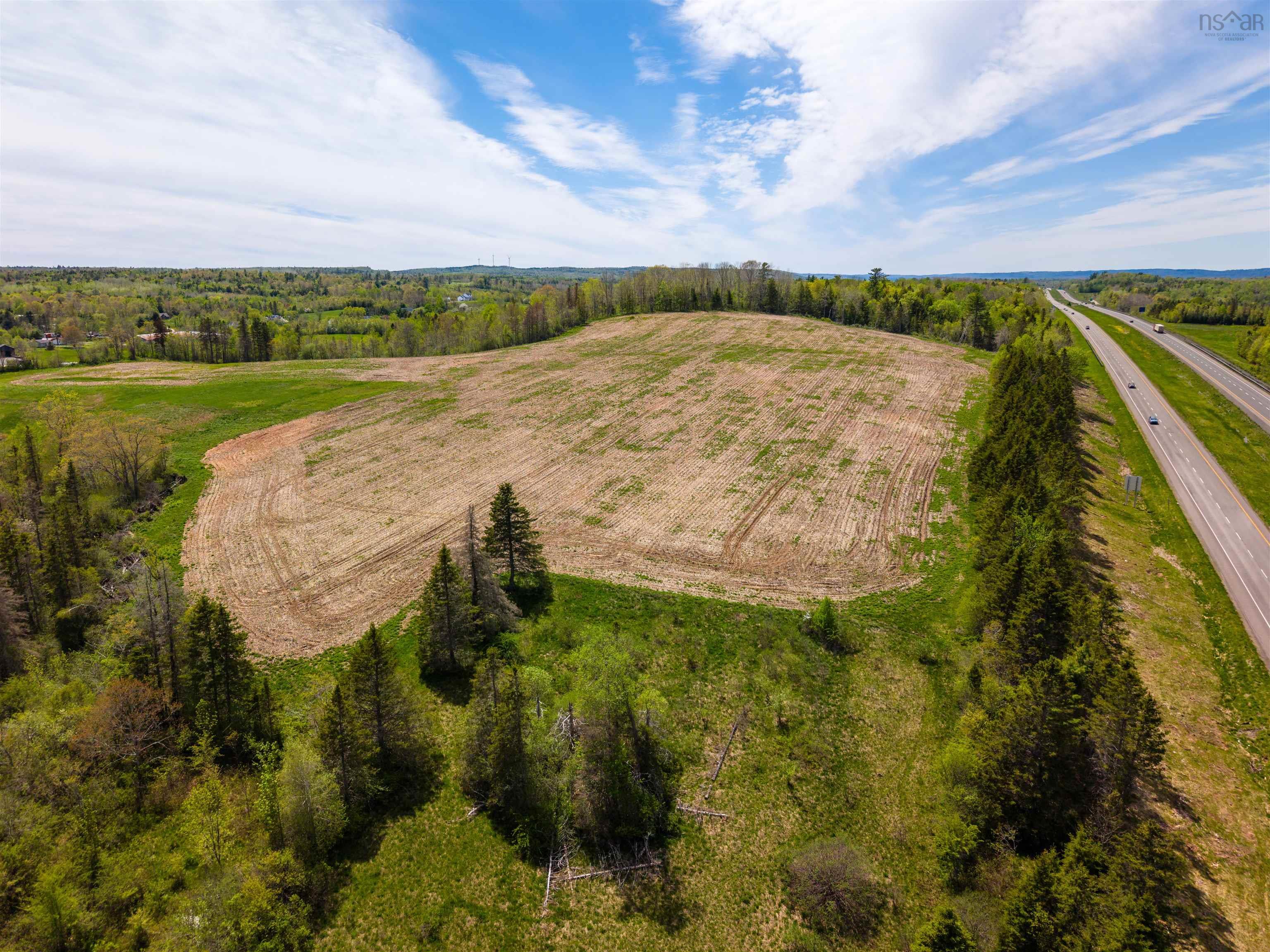 Aerial view of a large, flat, harvested field bordered by evergreen trees and a highway under a bright blue sky.