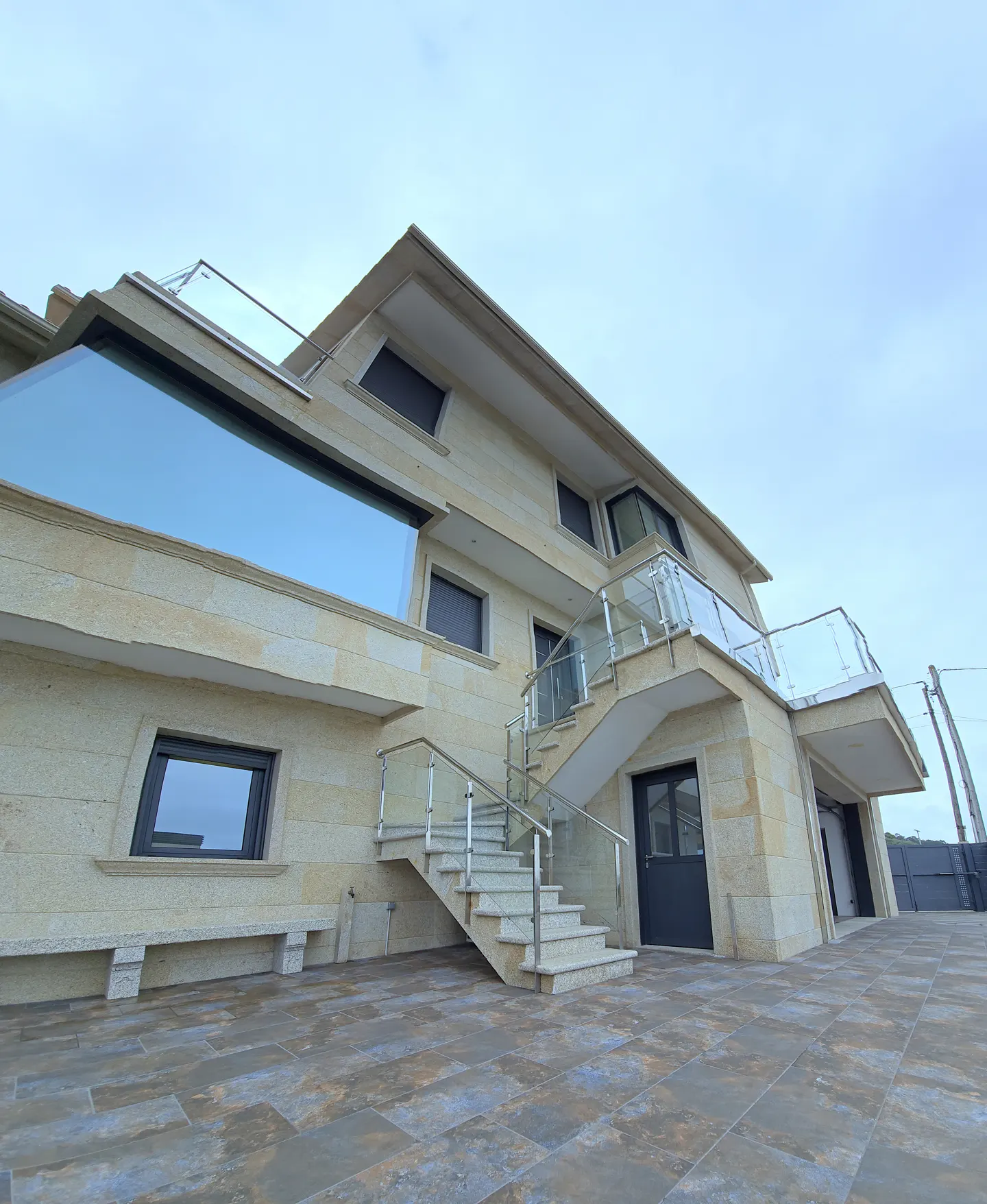Exterior view of a modern, multi-story beige stone house with glass railings and a staircase leading to a second-floor entrance.