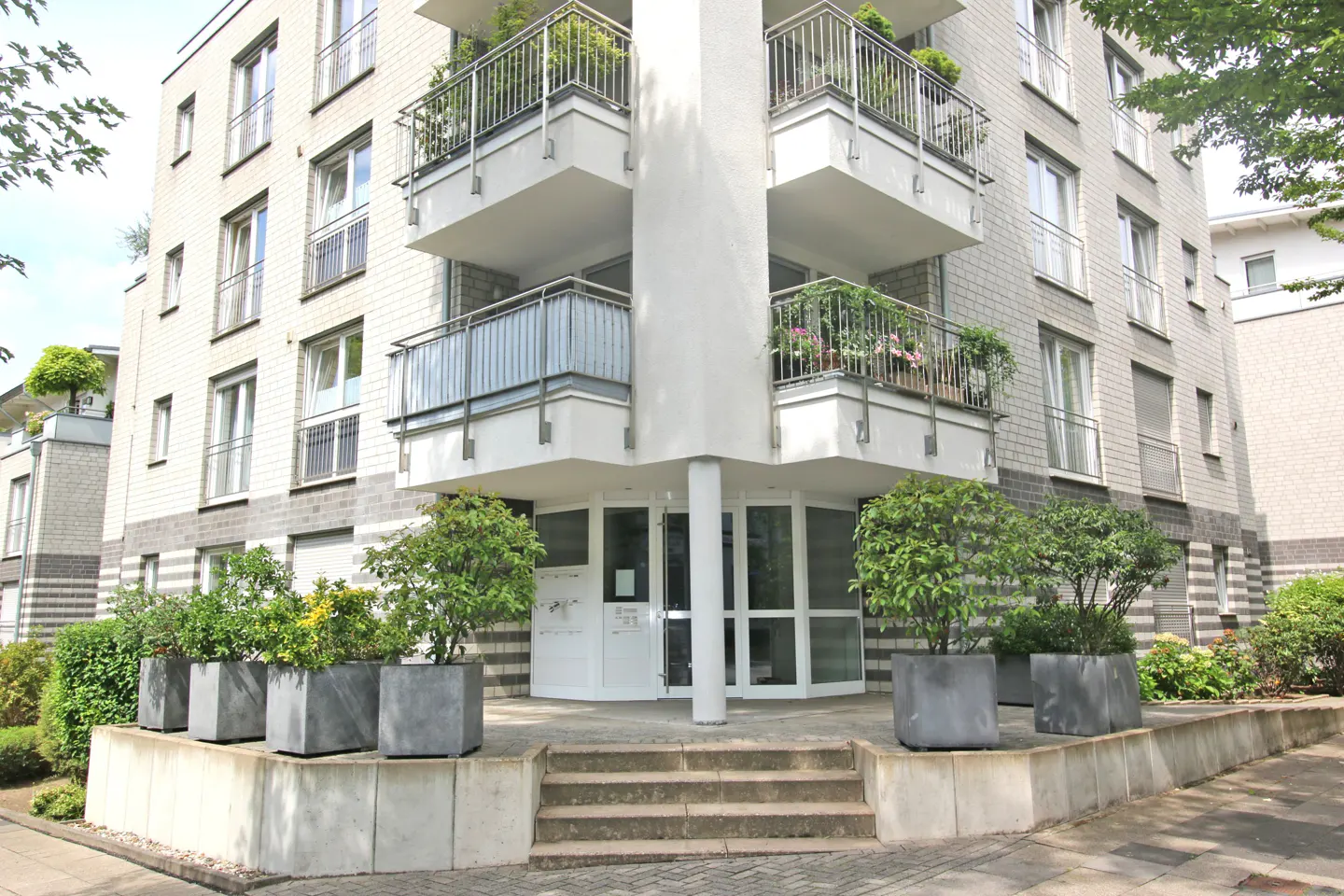Exterior of a modern apartment building with white brick, balconies, and potted plants at the entrance.
