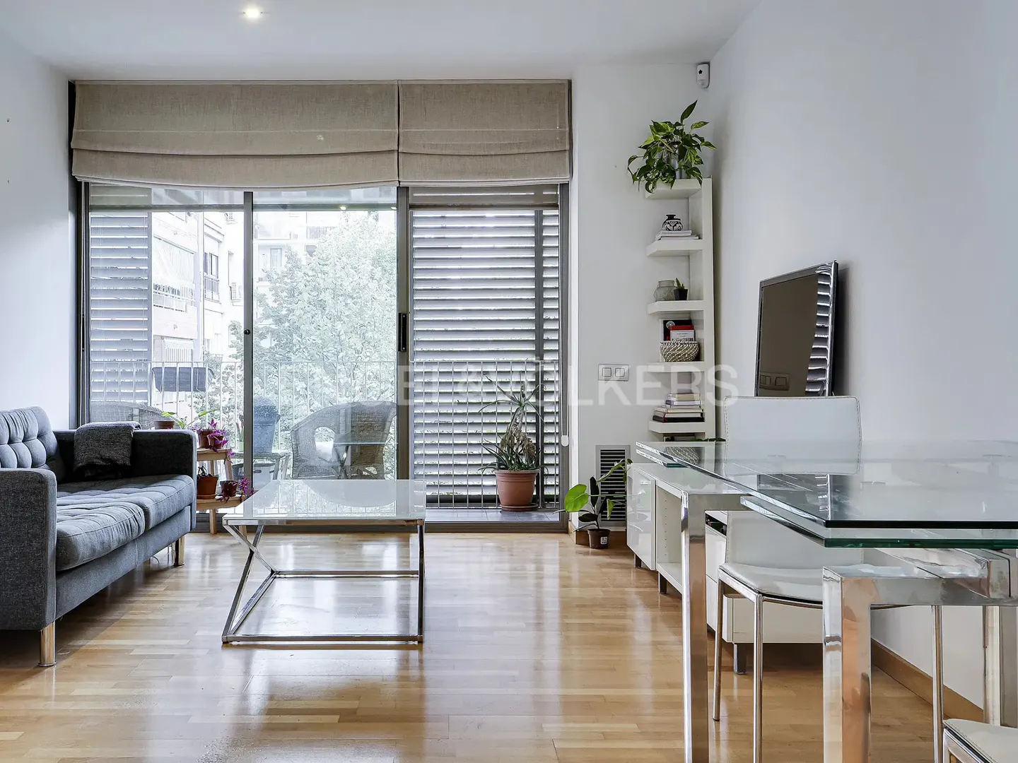 Bright living room with a gray sofa, glass coffee table, and dining set. Large windows with beige blinds overlook a balcony. Hardwood floors and white walls.