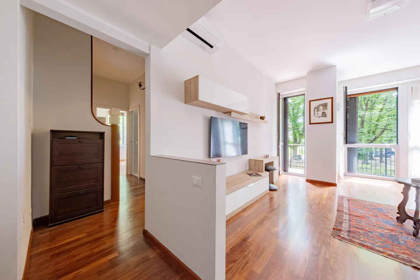 Modern living room with hardwood floors, wall-mounted TV, white media console, and a balcony view of trees.