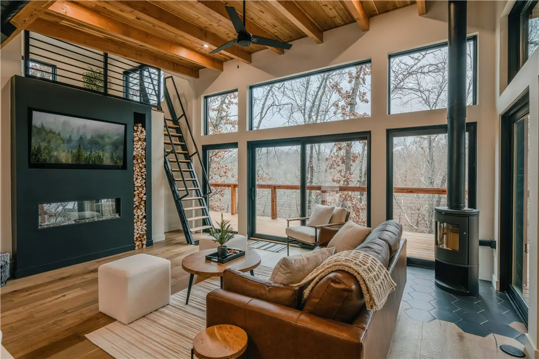 Living room with wood floors, a brown leather couch, and a black fireplace. Large windows overlook a wooded area.