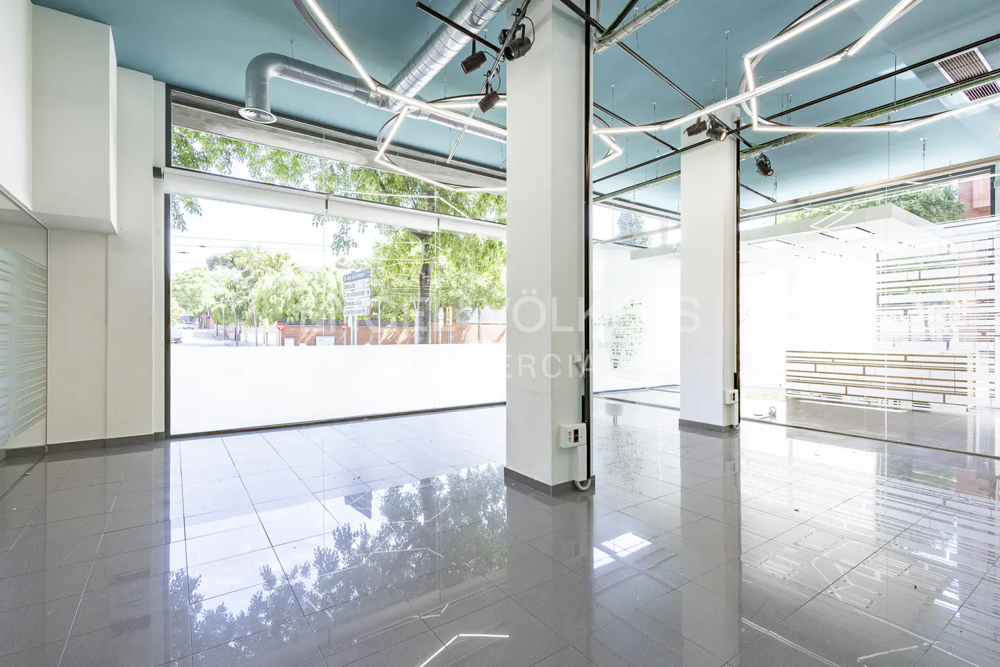 Empty commercial space with gray tile floors, white columns, and a blue ceiling with modern lighting. Large windows offer a street view.