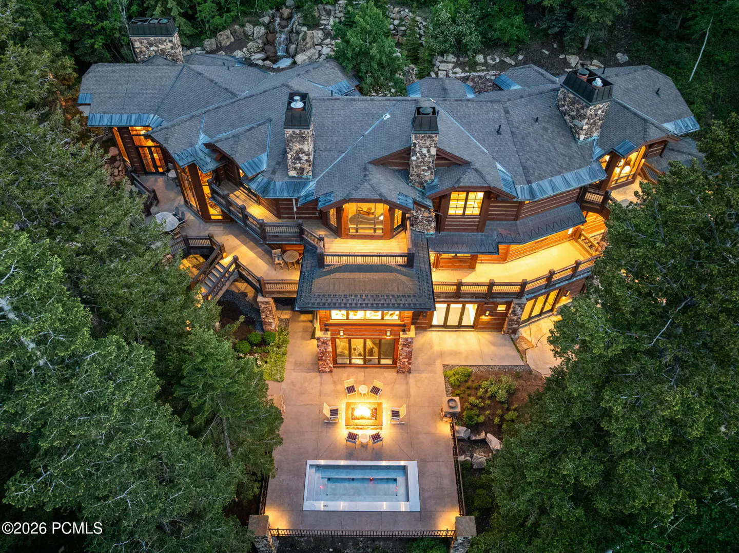 Aerial view of a large, multi-level log cabin home with a gray roof, stone chimneys, a pool, and a fire pit surrounded by trees.