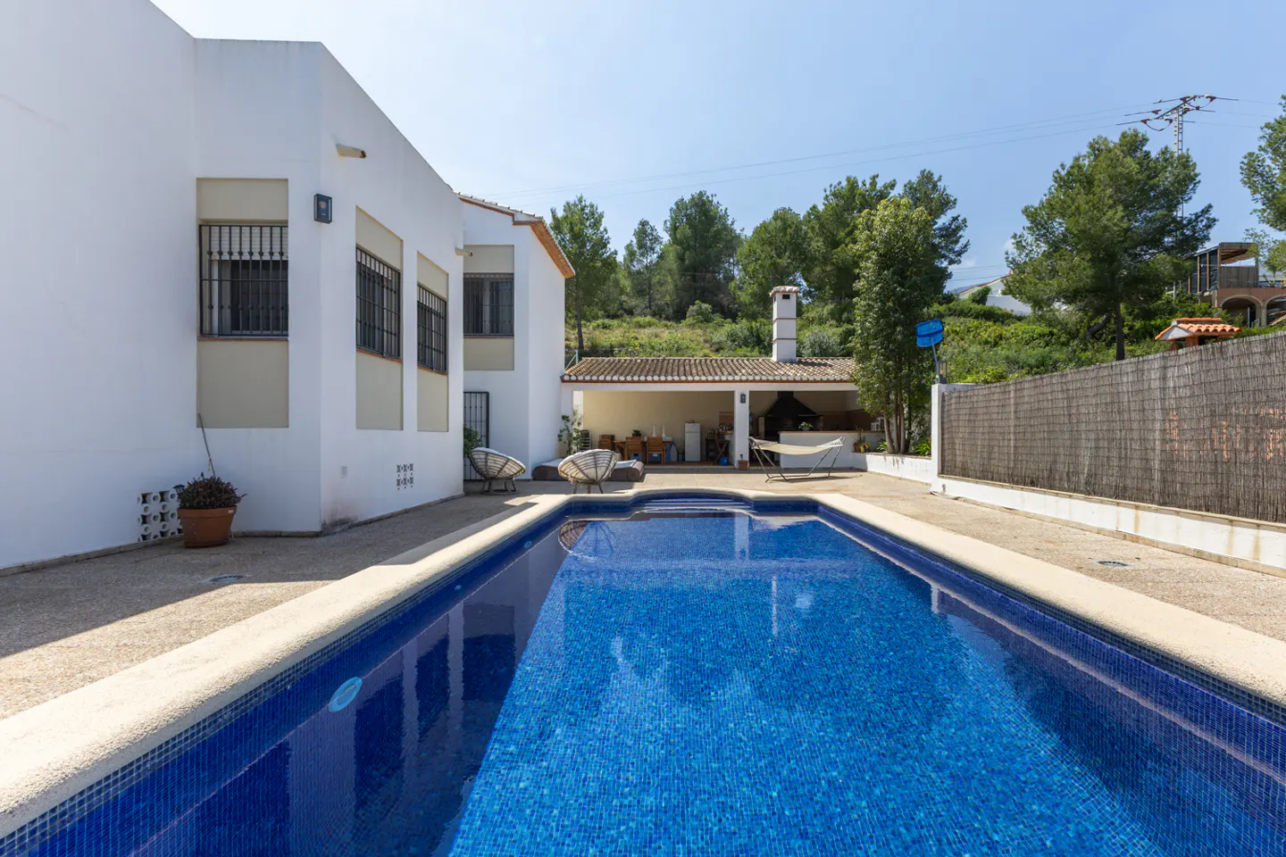 Outdoor pool with blue tiles in a backyard. White house with black barred windows, patio with hammock, and green trees in the background.