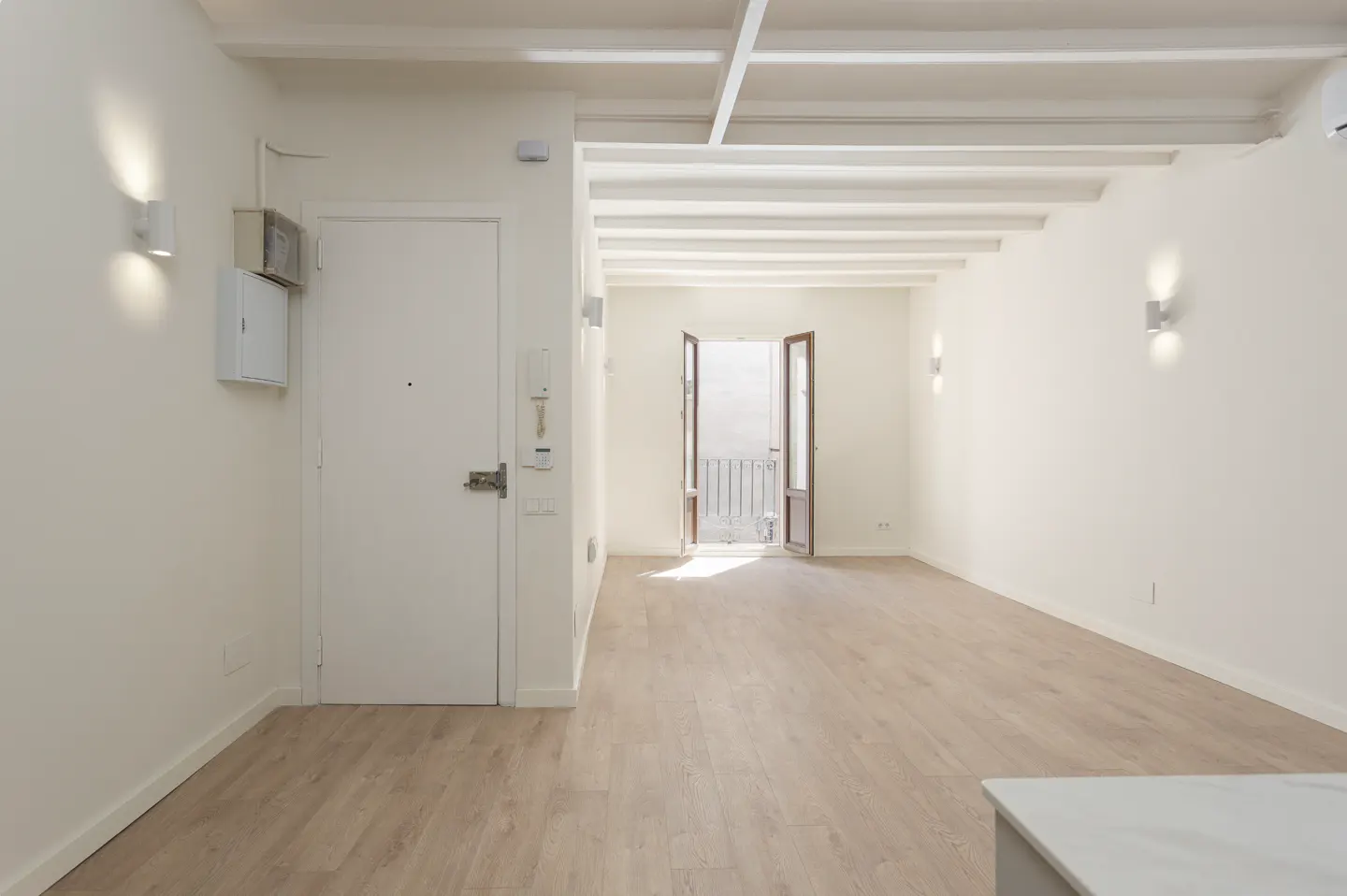 Minimalist white room with light wood floors, exposed ceiling beams, wall sconces, and an open door to a small balcony.