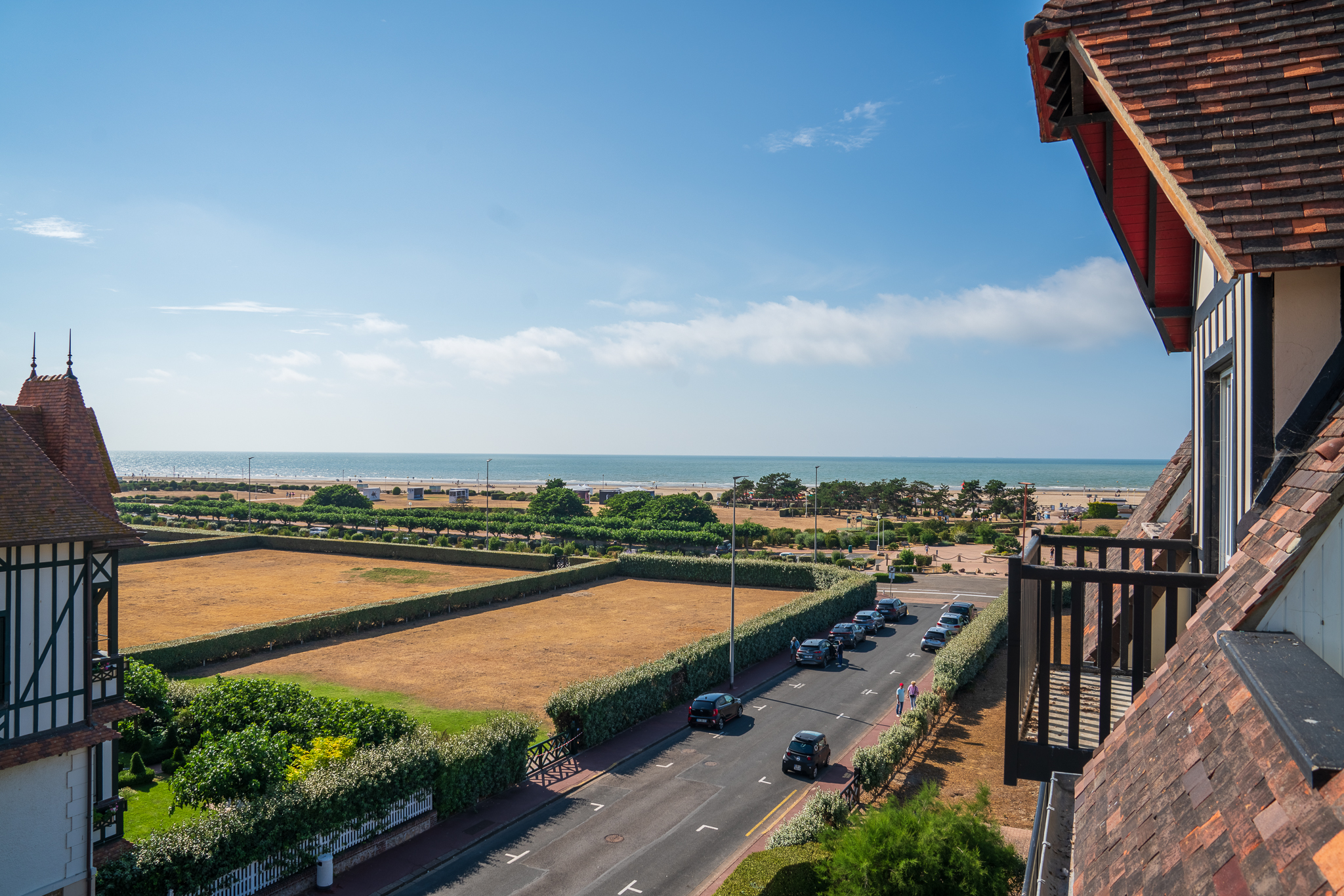High-angle view of a coastal street with parked cars, a dry field, and a sandy beach under a clear blue sky.