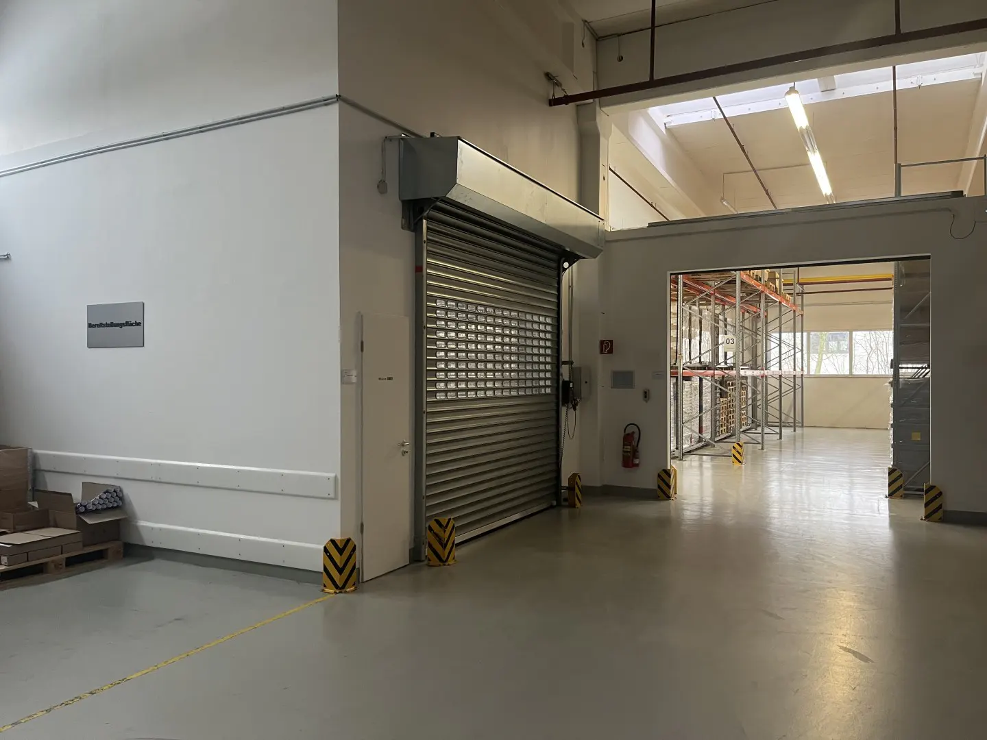 Interior view of a warehouse with a metal roll-up door, gray floor, white walls, and shelving visible through an open doorway.