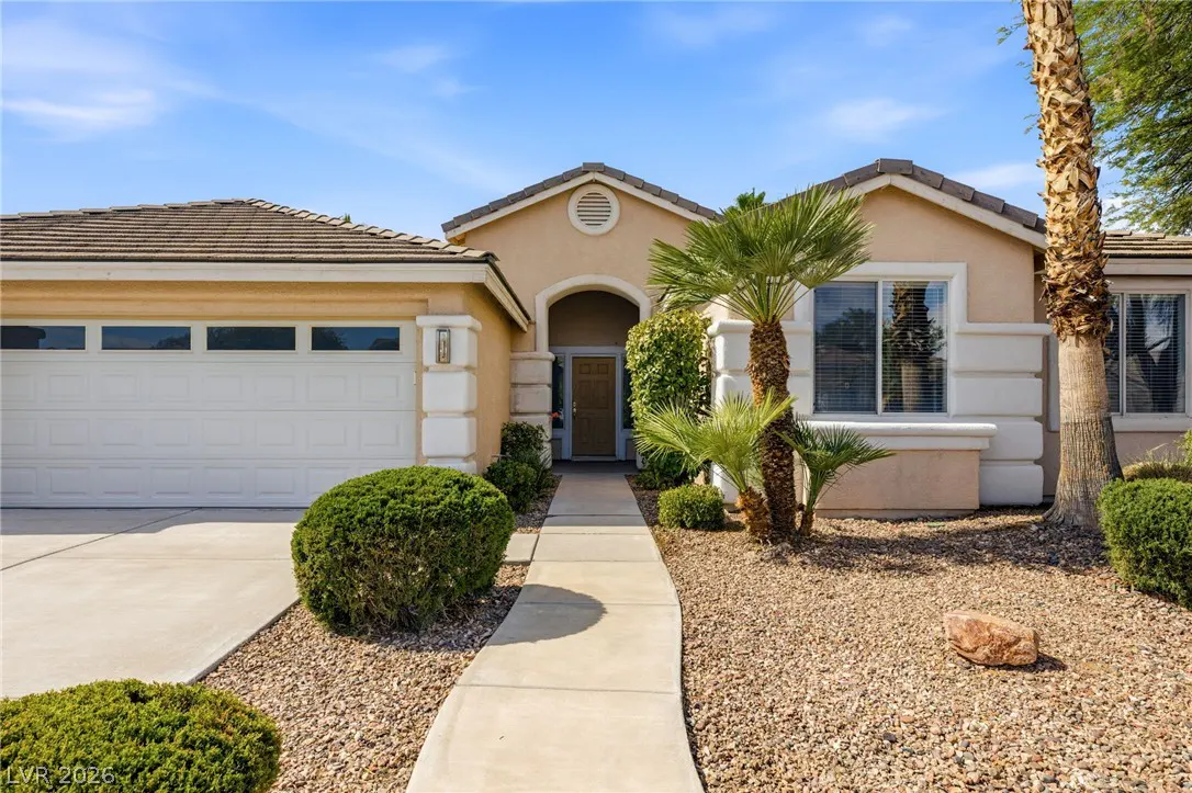 Single-story tan stucco home with a white garage, desert landscaping, palm tree, and a concrete walkway to the front door.