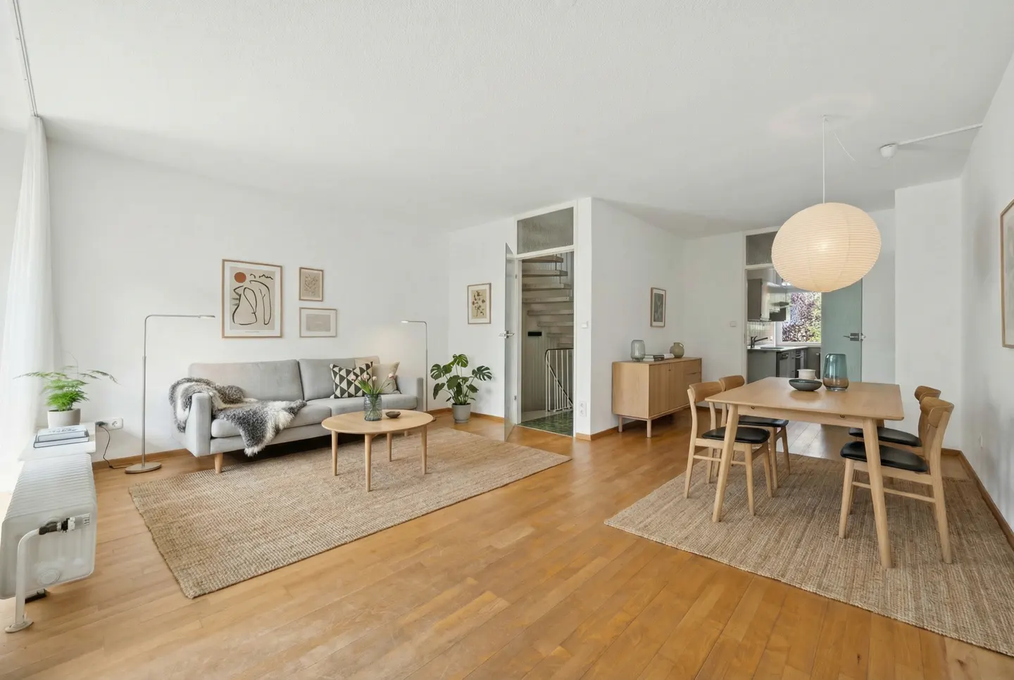 Bright, open-plan living and dining area with hardwood floors. A gray sofa, wood table, and dining set sit on jute rugs. White walls and natural light.