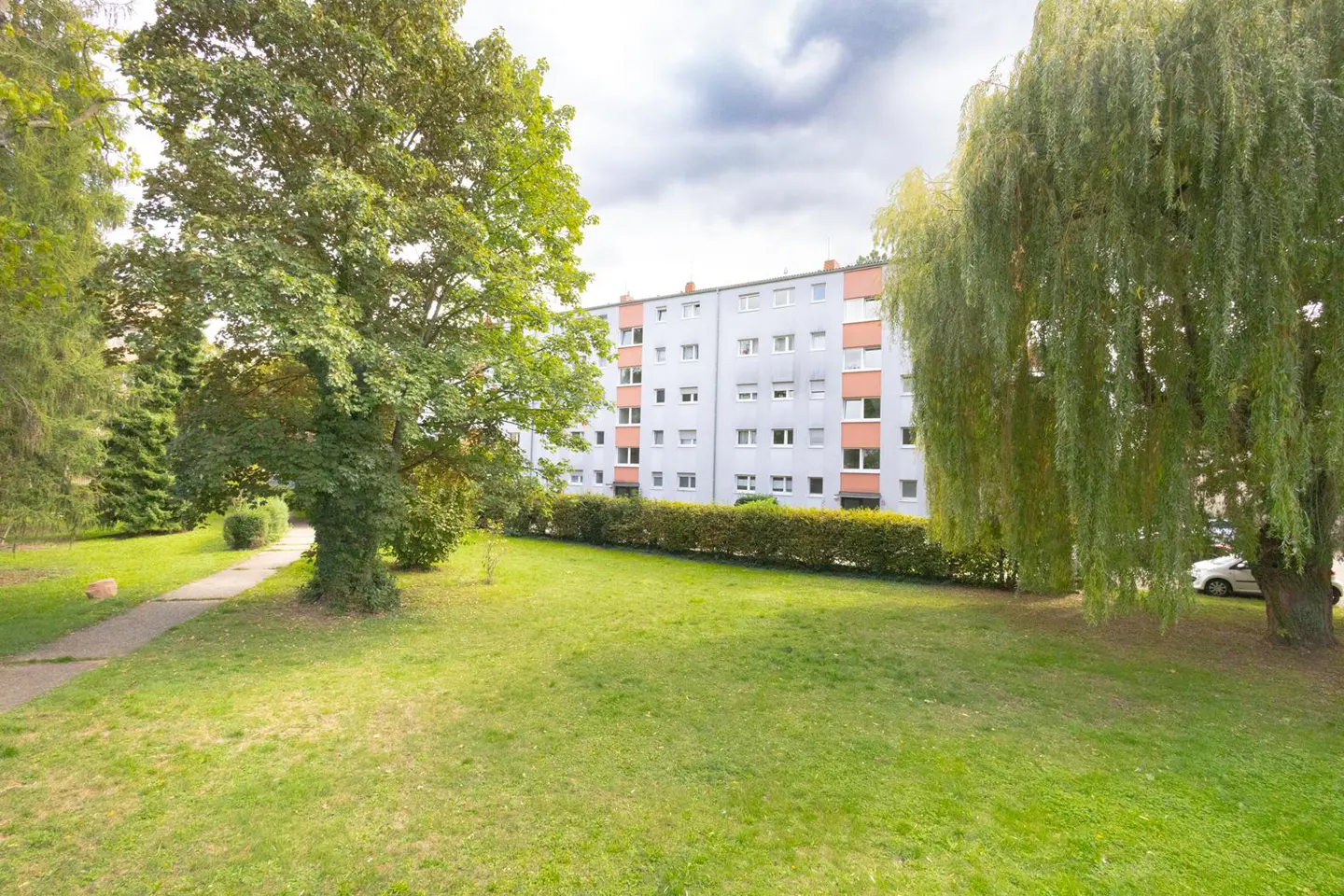 Apartment building with green lawn, trees, and cloudy sky. The building is light gray with orange accents. A sidewalk leads to the left.