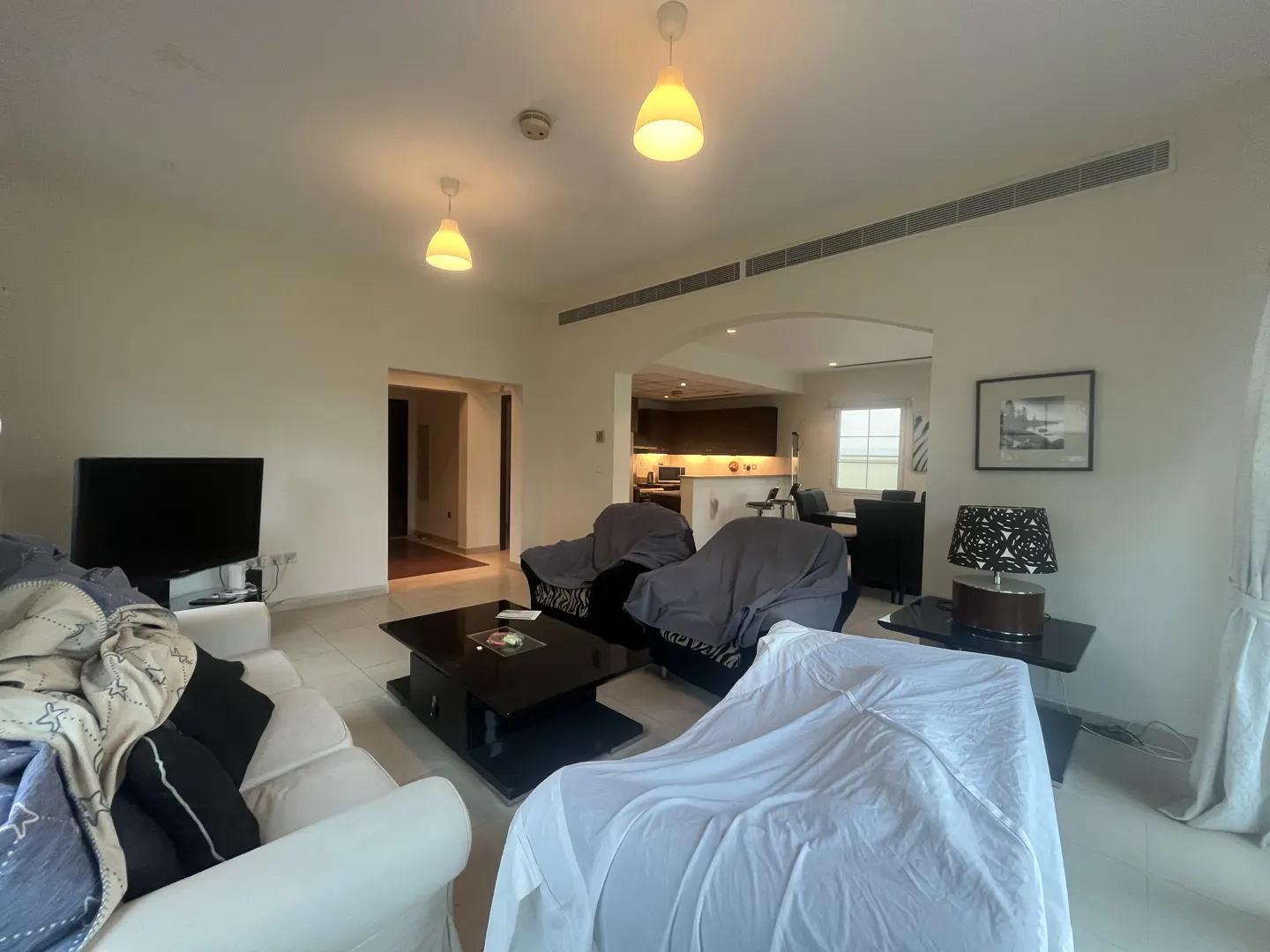 A bright living room with furniture covered in sheets, a black TV, and a view into the kitchen.