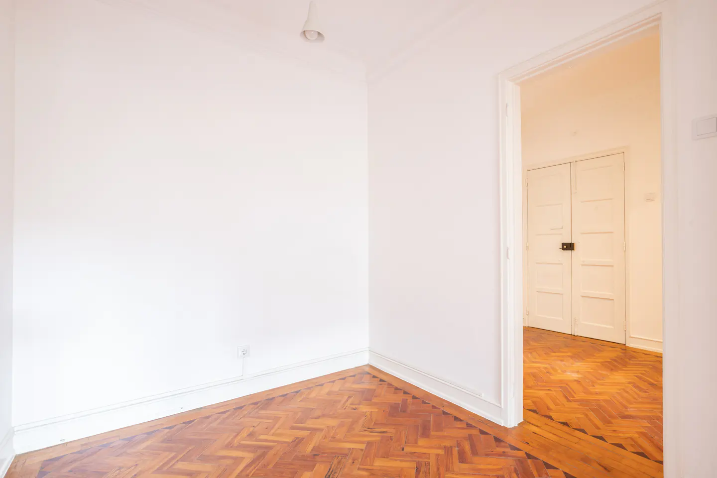 Empty room with white walls, herringbone wood floor, and open doorway to another room with double doors.