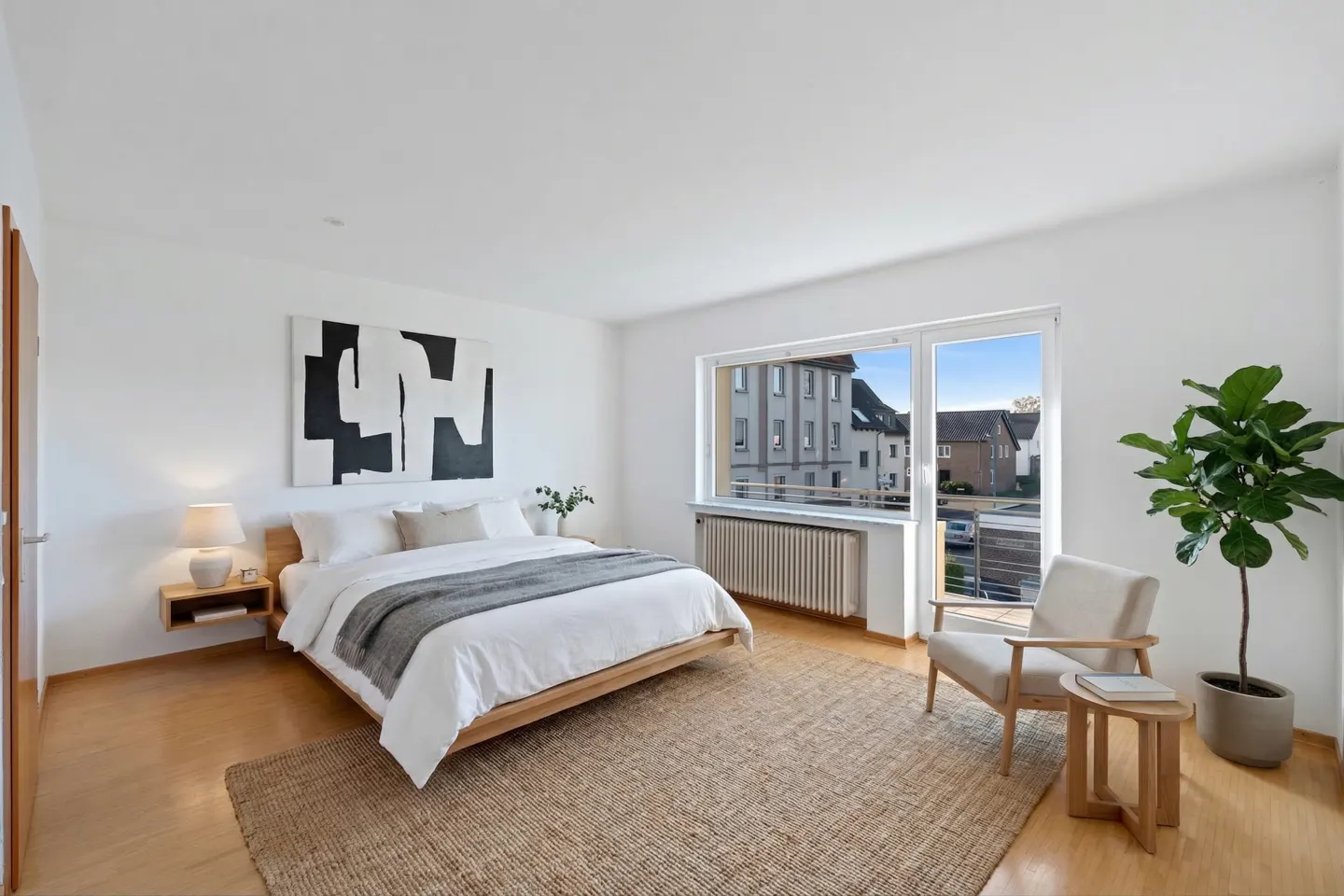Bright bedroom with white walls, wood floors, and a large window. A bed with white linens and a gray throw sits beneath a black and white abstract painting. A jute rug and armchair complete the space.