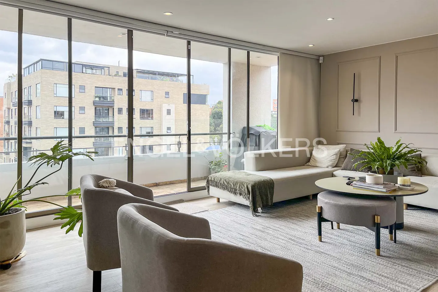 Living room with two gray chairs, a white sofa, and a round table on a gray rug. Large windows overlook a city building.