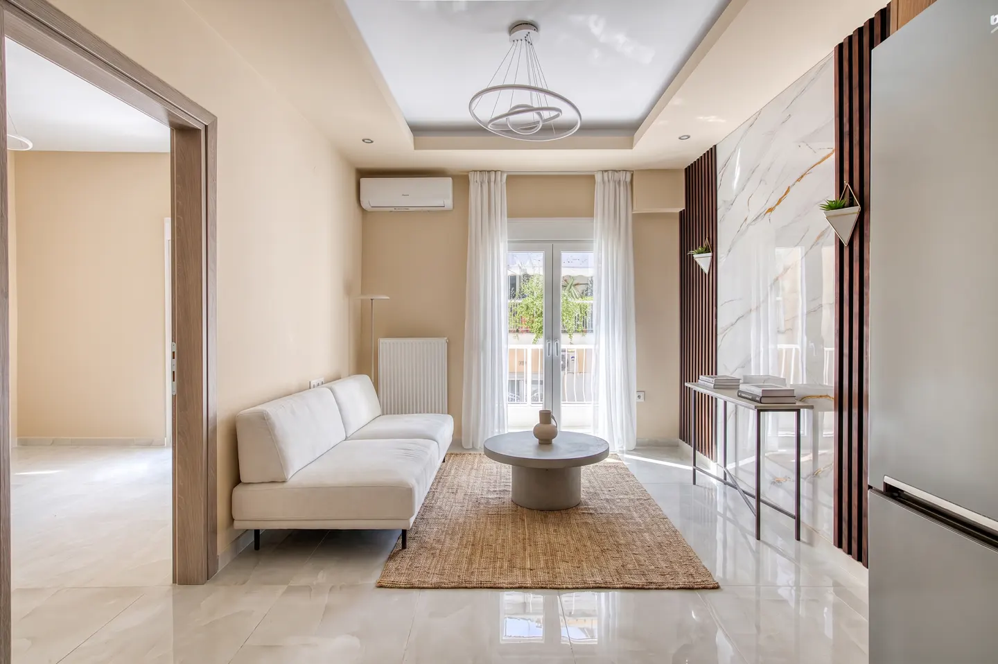 Living room with white sofa, round table on jute rug, and marble accent wall with wood slats. Light beige walls and white curtains.
