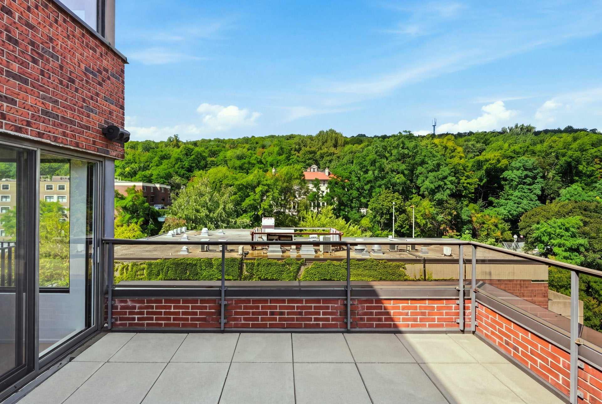 A balcony with red brick walls and gray tile flooring overlooking a lush green forest and distant buildings under a blue sky. [AI-generated text]