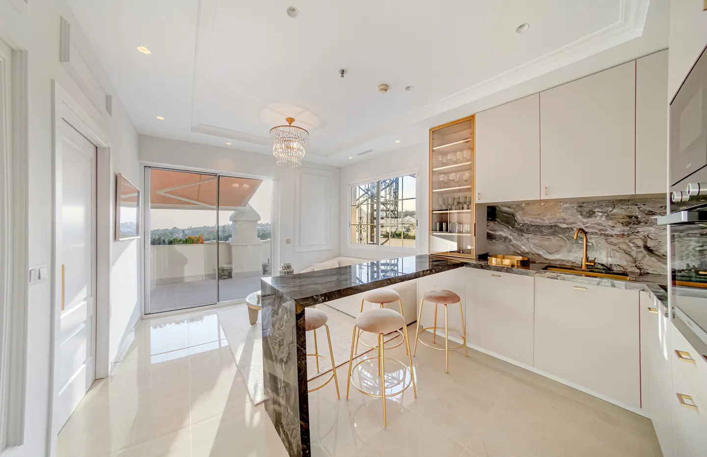 Bright, modern kitchen with white cabinets, marble counters, and gold accents. Stools line a black marble island. A chandelier hangs above.