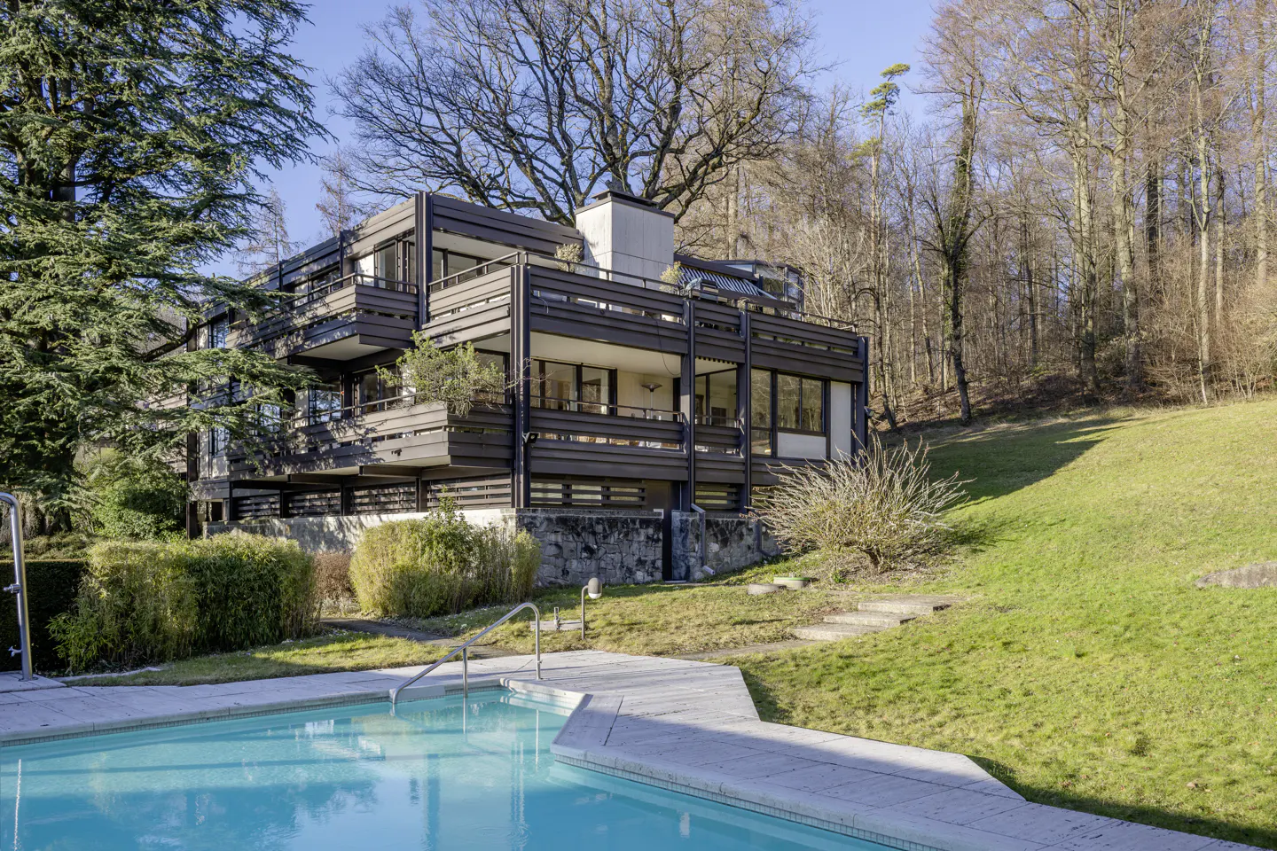 Modern brown apartment building with balconies overlooks a turquoise swimming pool and grassy hillside.