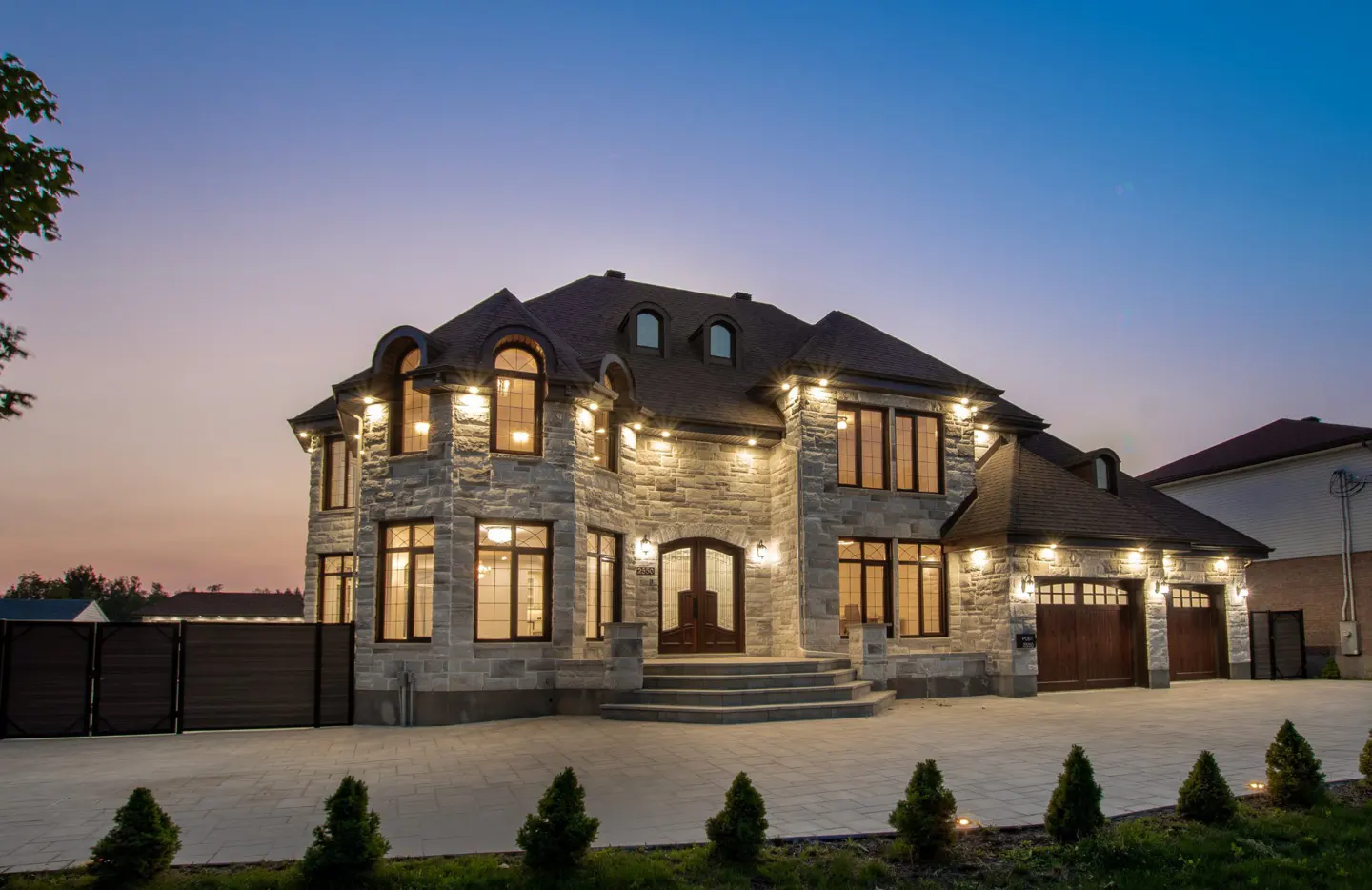 Exterior view of a large, two-story stone house with a dark roof and a three-car garage at dusk.