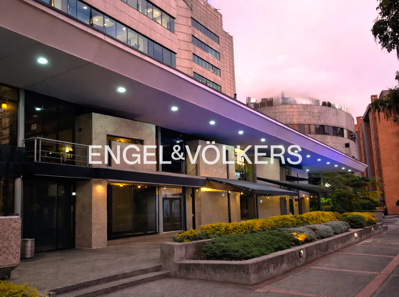 Engel & Völkers office exterior at dusk. Modern building with a long, lit awning and manicured landscaping.