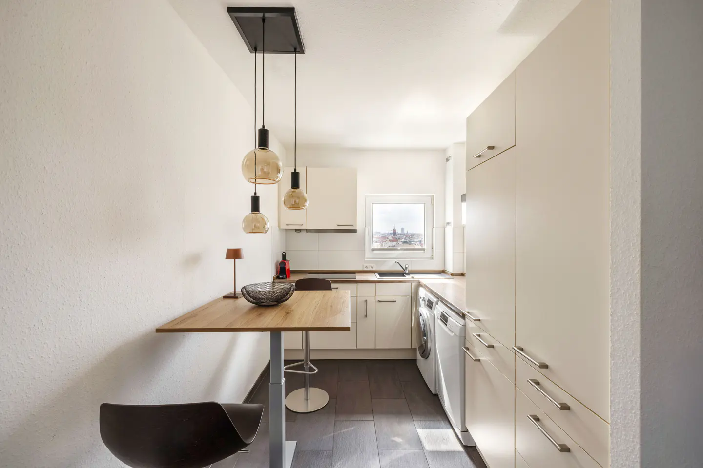 Bright, modern kitchen with white cabinets, wood countertop, and pendant lights. A window shows a city view.