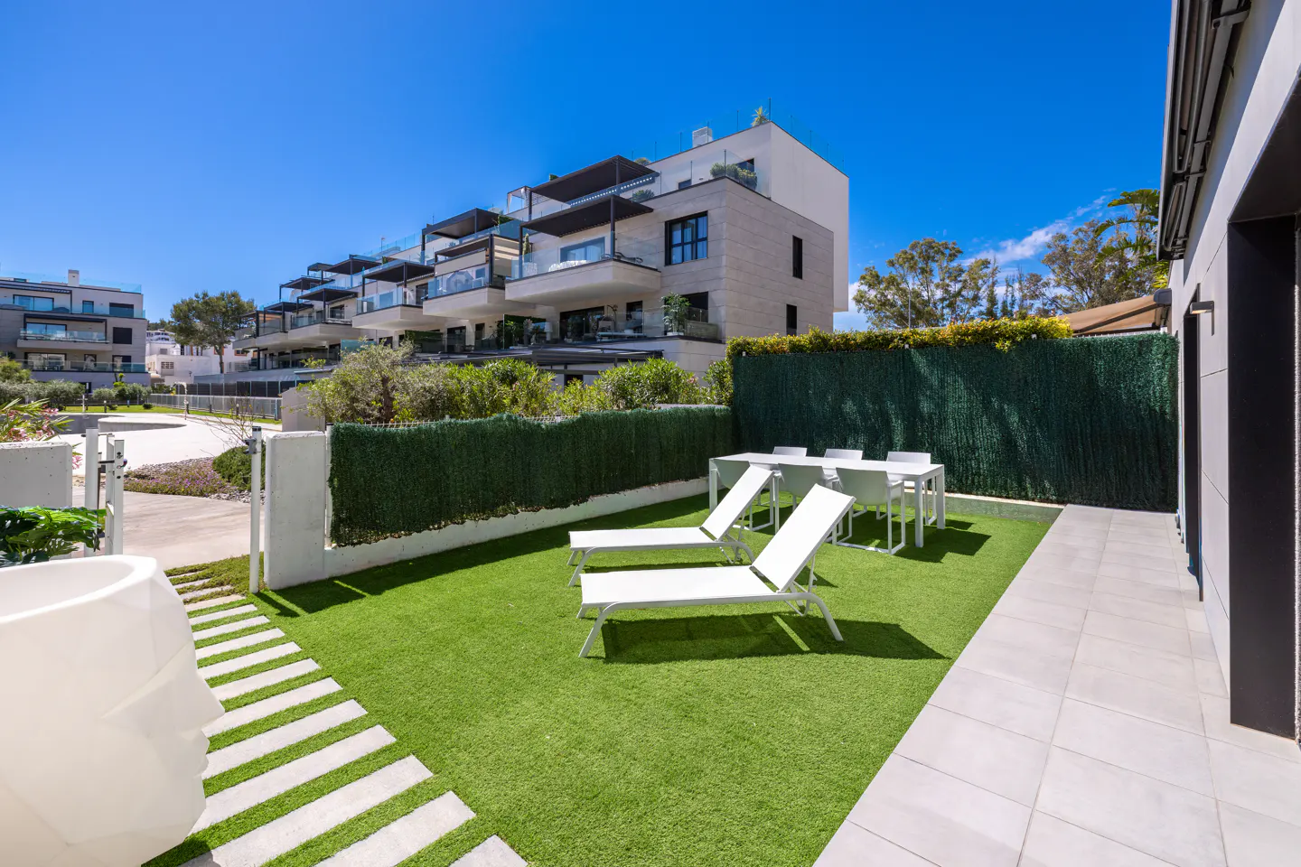 Backyard with green grass, white lounge chairs, and a table with chairs. Modern apartment buildings are in the background.