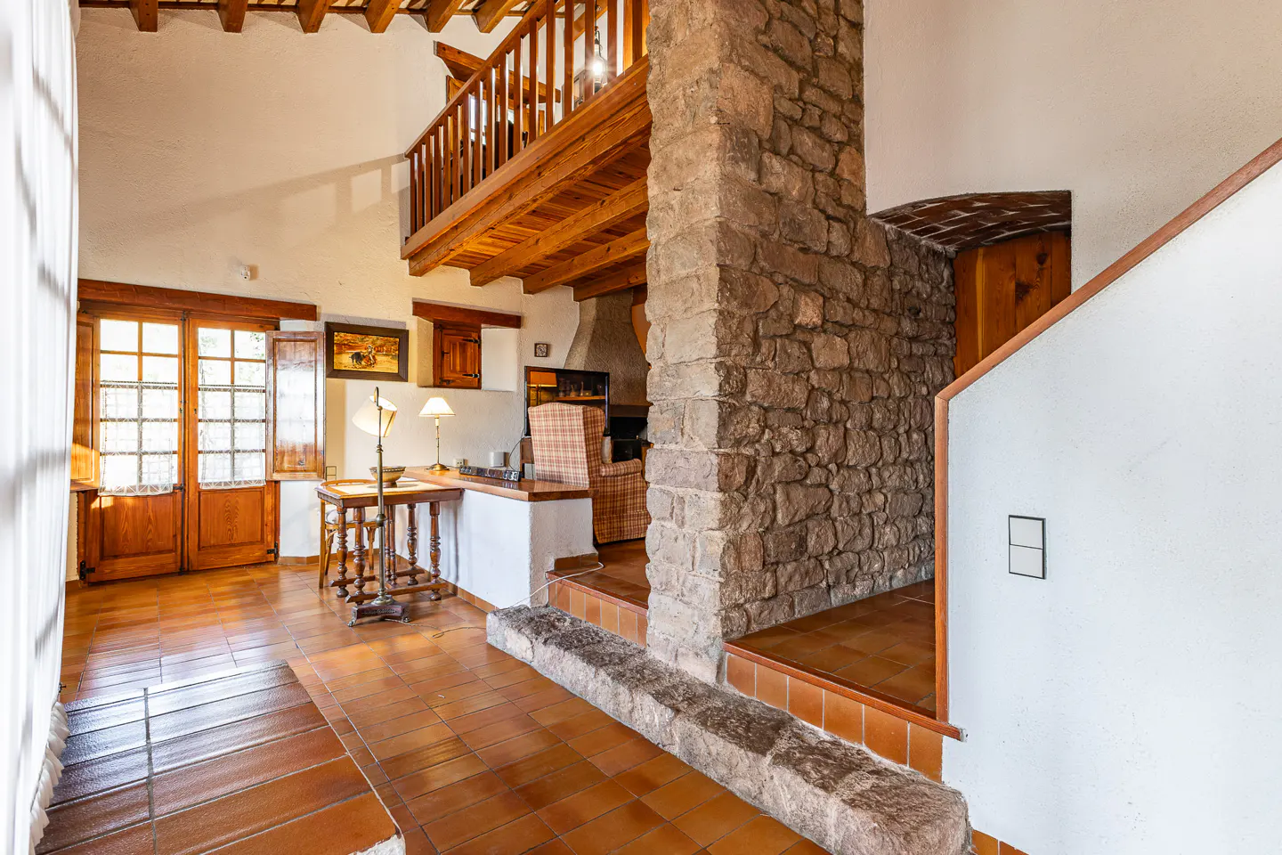 Interior view of a home with terracotta tile floors, a stone wall, and a wooden balcony. Natural light streams through the windows.