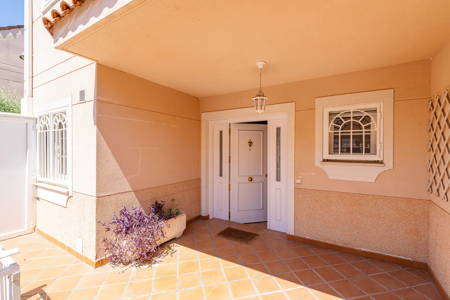 A beige house entrance with a white door, tiled porch, a hanging lantern, and a planter with purple foliage.