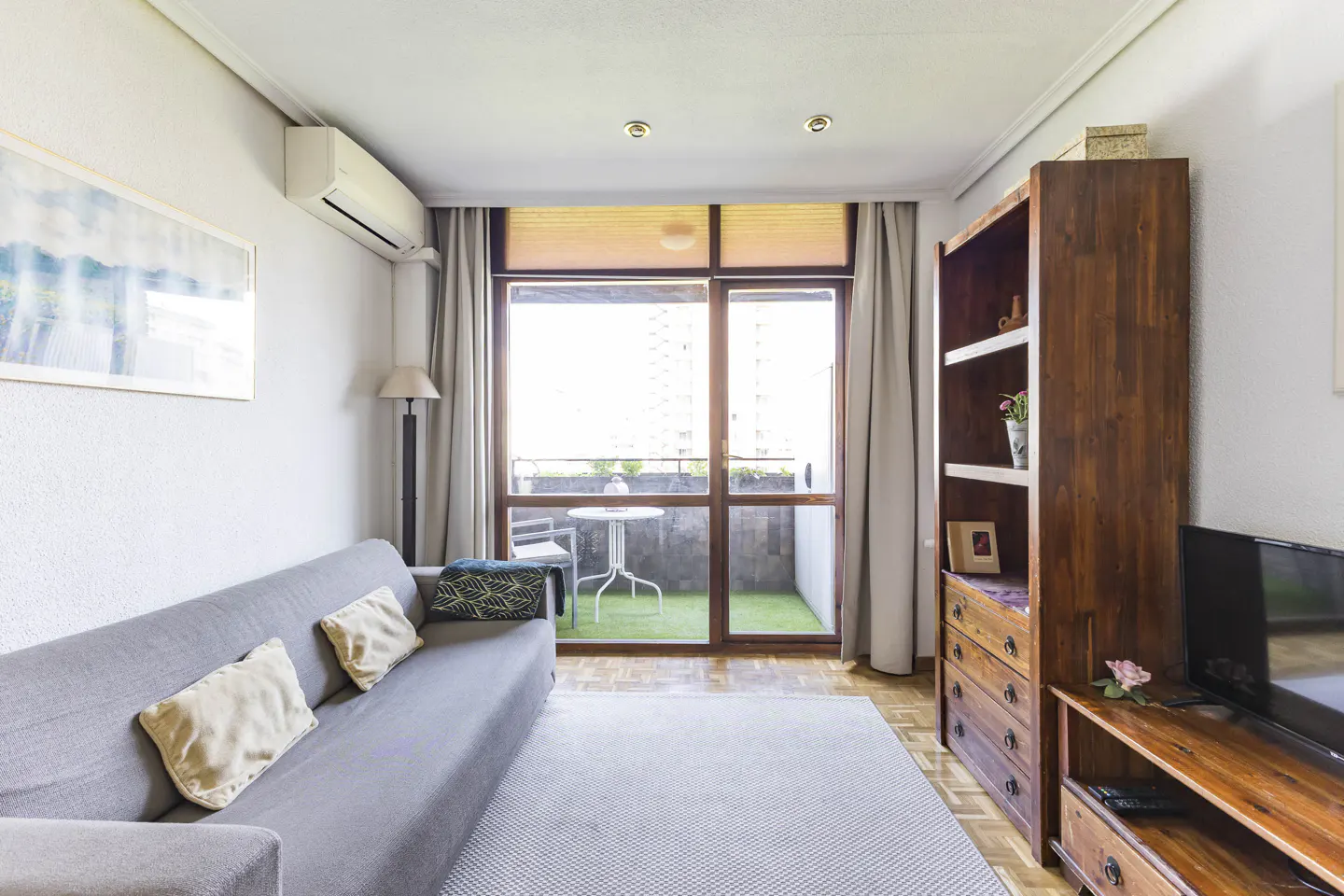 Living room with gray sofa, wood bookcase, and balcony with table and chairs. Neutral colors and natural light.