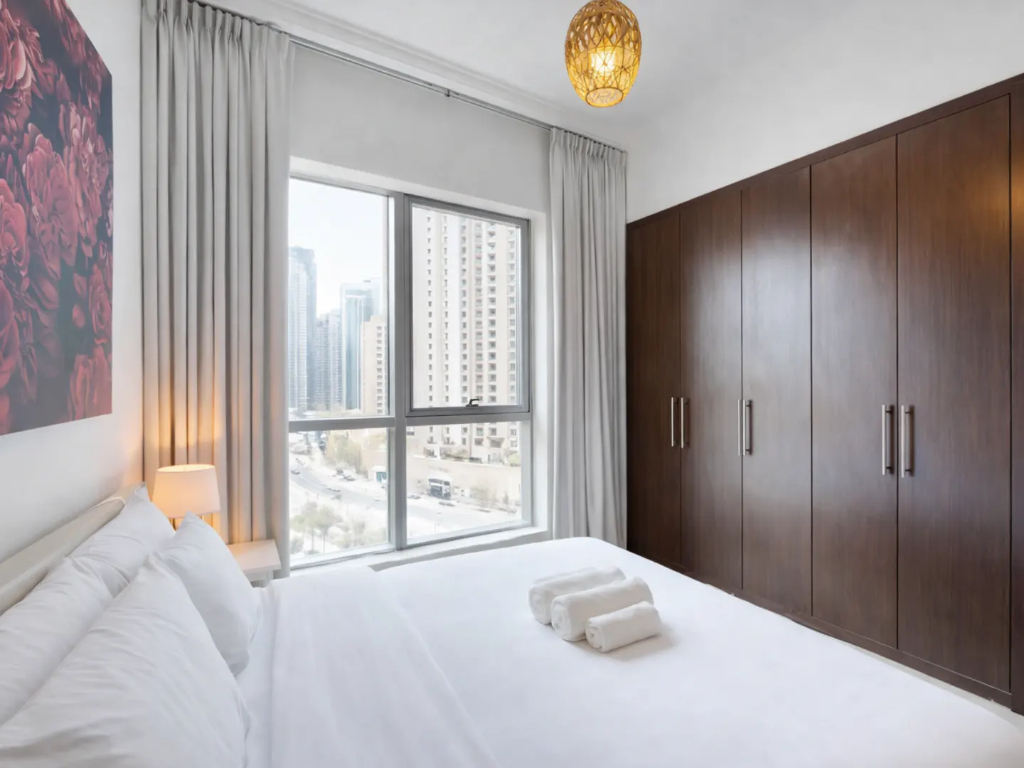 Bedroom with white linens, rolled towels, and a city view. Brown closet doors and a woven light fixture add warmth. Floral art on the wall.