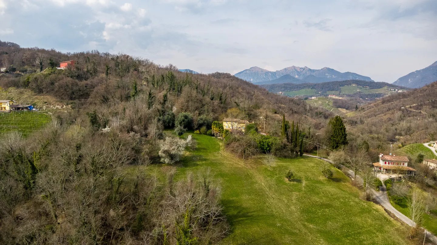 Hilly landscape with green fields, bare trees, and scattered houses. Mountains are visible in the background under a cloudy sky.