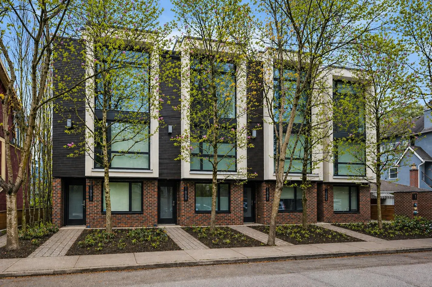 Three modern townhouses with black siding, brick bases, and large windows, framed by young trees.