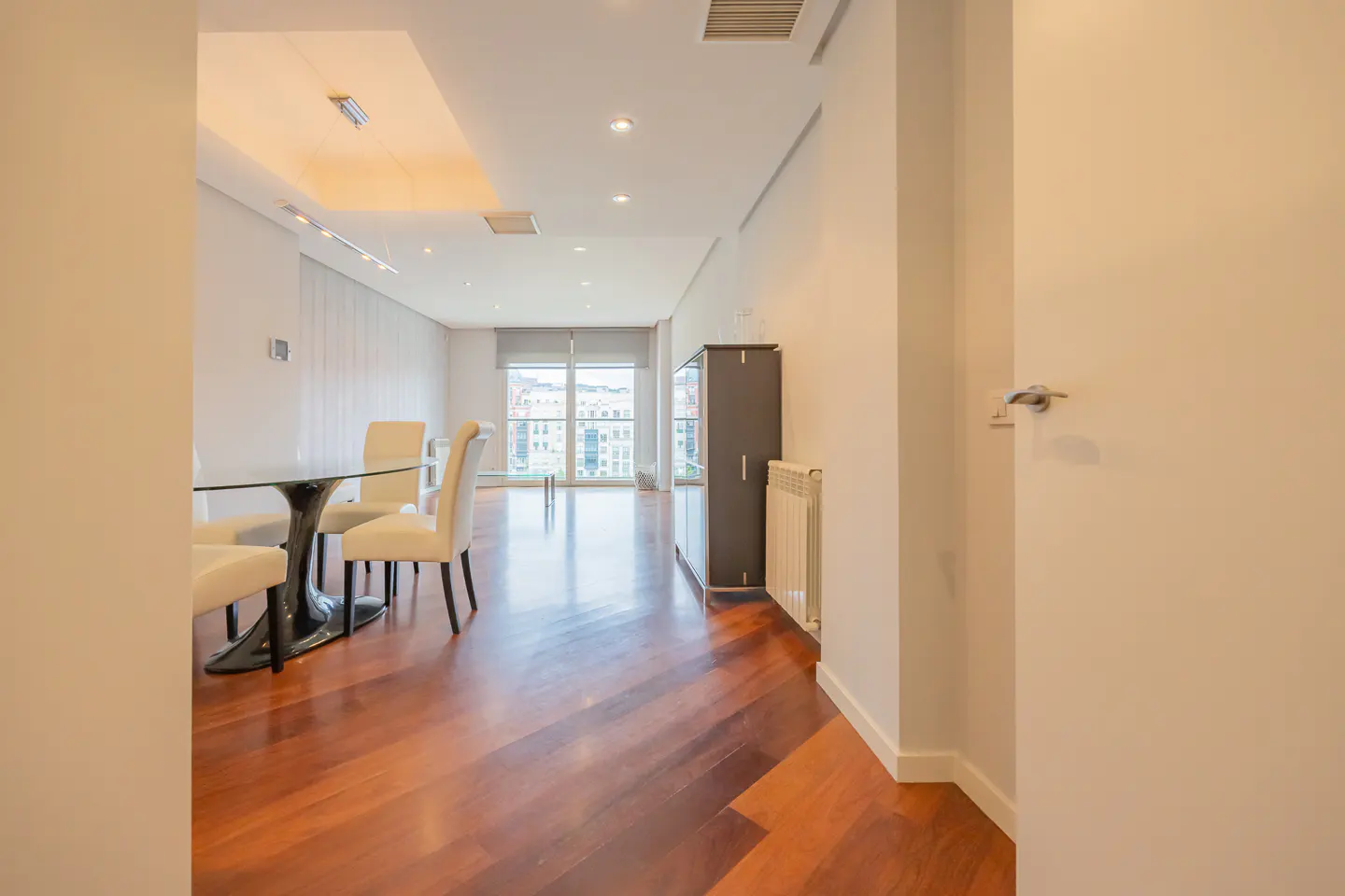 Interior view of a modern apartment with hardwood floors, a glass dining table with cream chairs, and a large window overlooking a city.