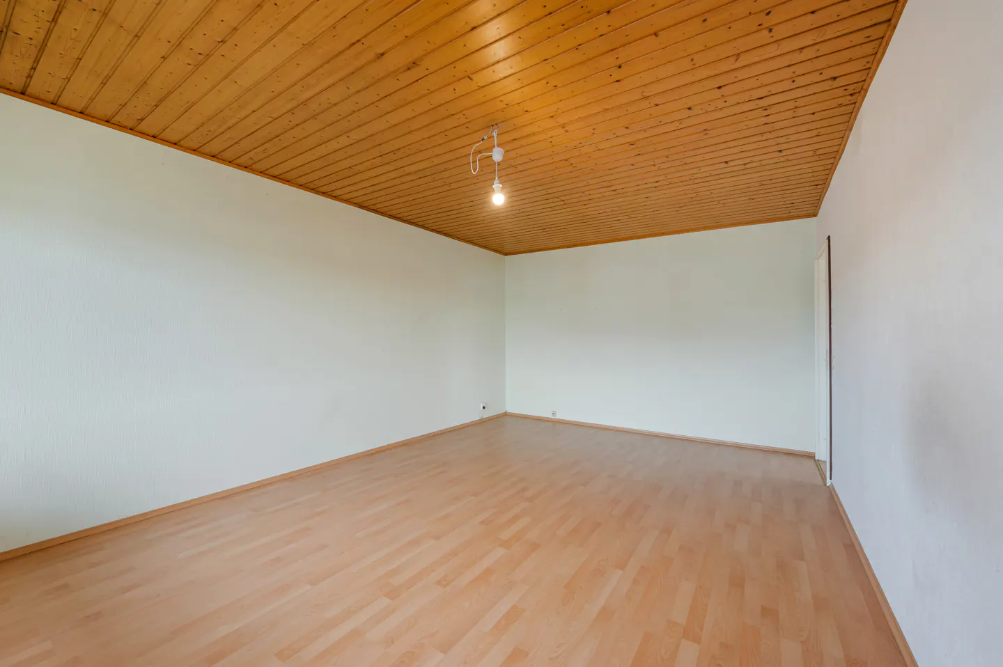 Empty room with light wood floor, white walls, and wood plank ceiling. A single light bulb hangs from the ceiling.