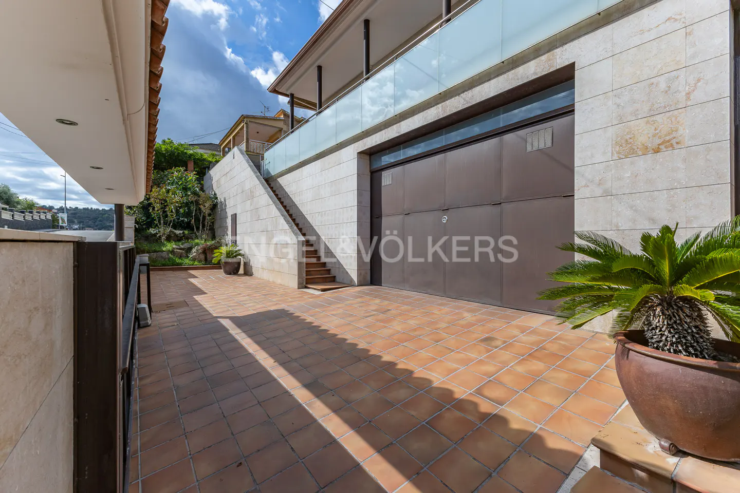 Exterior view of a house with a brown garage door, stone walls, and a tiled driveway. Stairs lead to a balcony with a glass railing. A potted palm sits on the right.