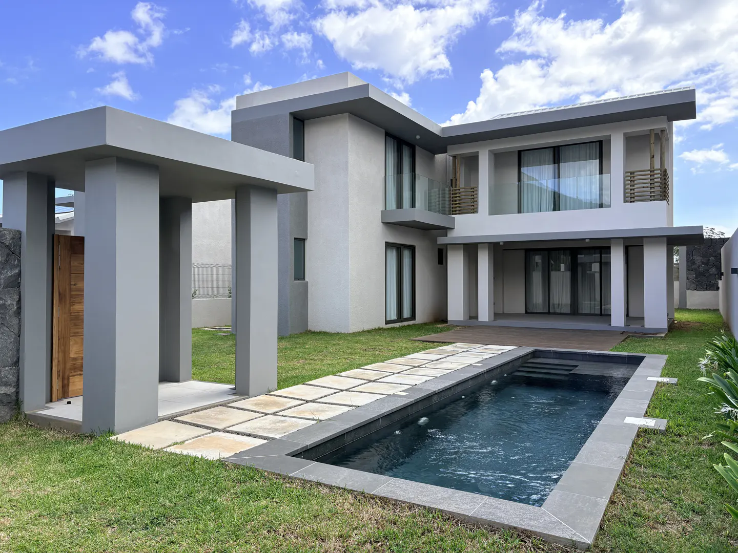 Two-story modern house with a gray exterior, a pool, and a stone walkway on a green lawn under a blue sky.
