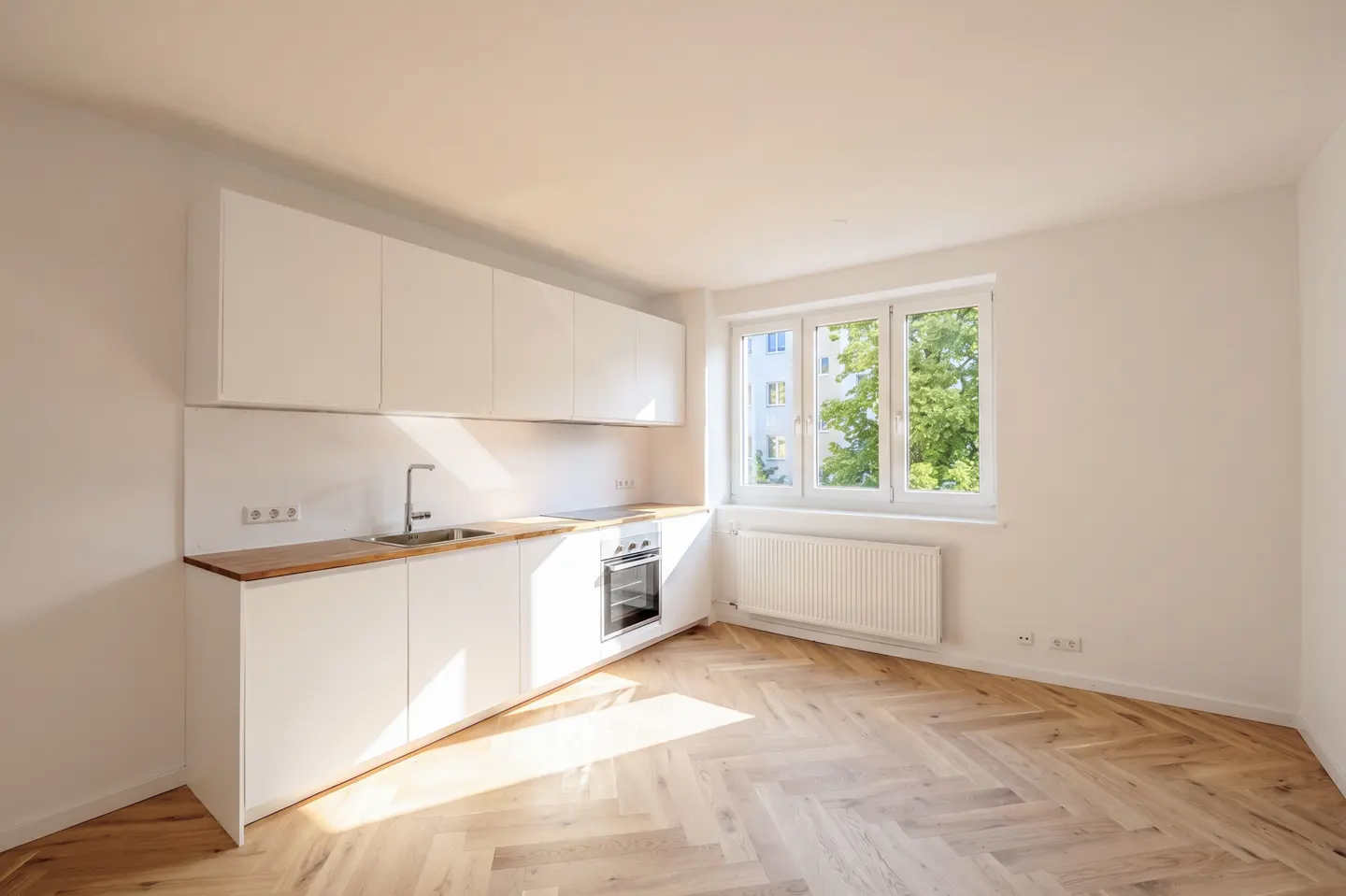 Bright, modern kitchen with white cabinets, wood countertop, and herringbone wood floor. A window overlooks green trees.