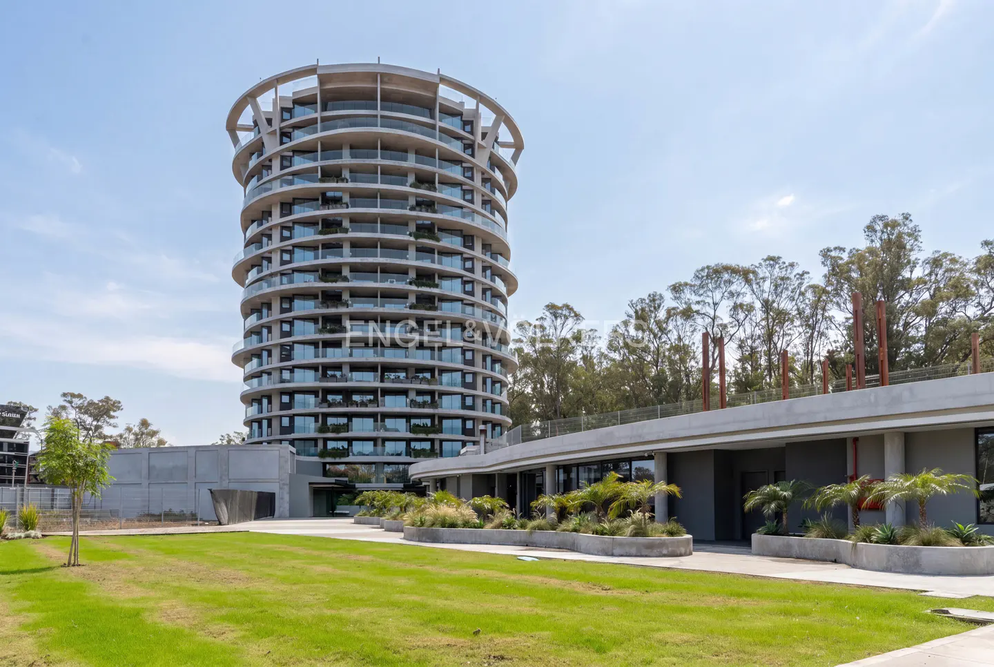 A modern, cylindrical high-rise apartment building with glass balconies stands next to a low-rise structure and lawn.