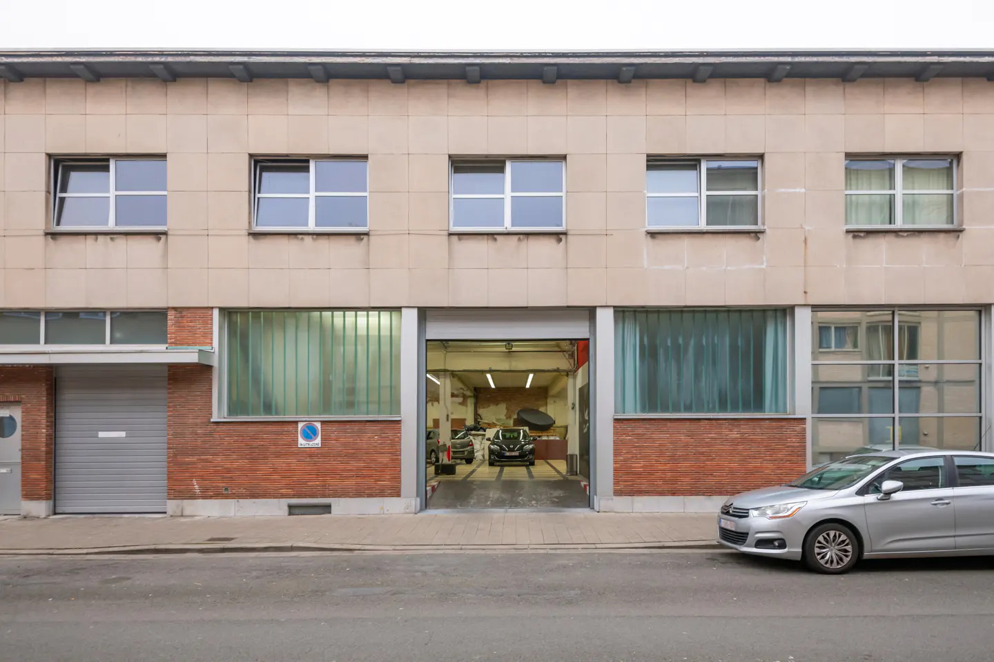 Two-story beige building with a garage door open to a car repair shop. A silver car is parked on the street.