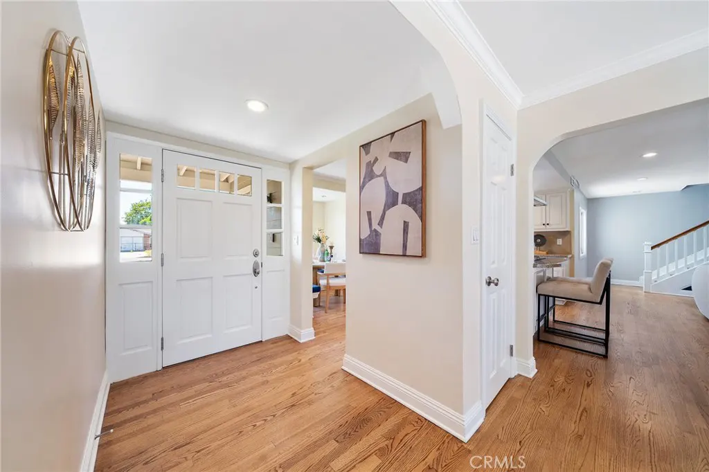 A bright foyer with hardwood floors, white walls, and a white front door with glass panels. An abstract art piece hangs on the wall.