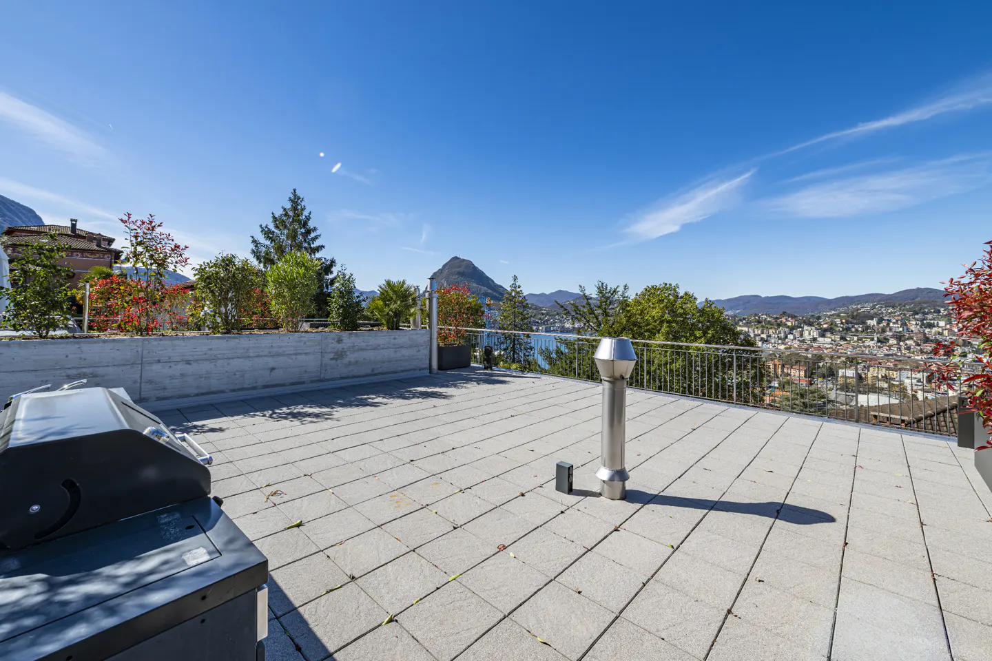 Rooftop patio with grill, gray pavers, and metal railing overlooking a city, lake, and mountains under a clear blue sky.